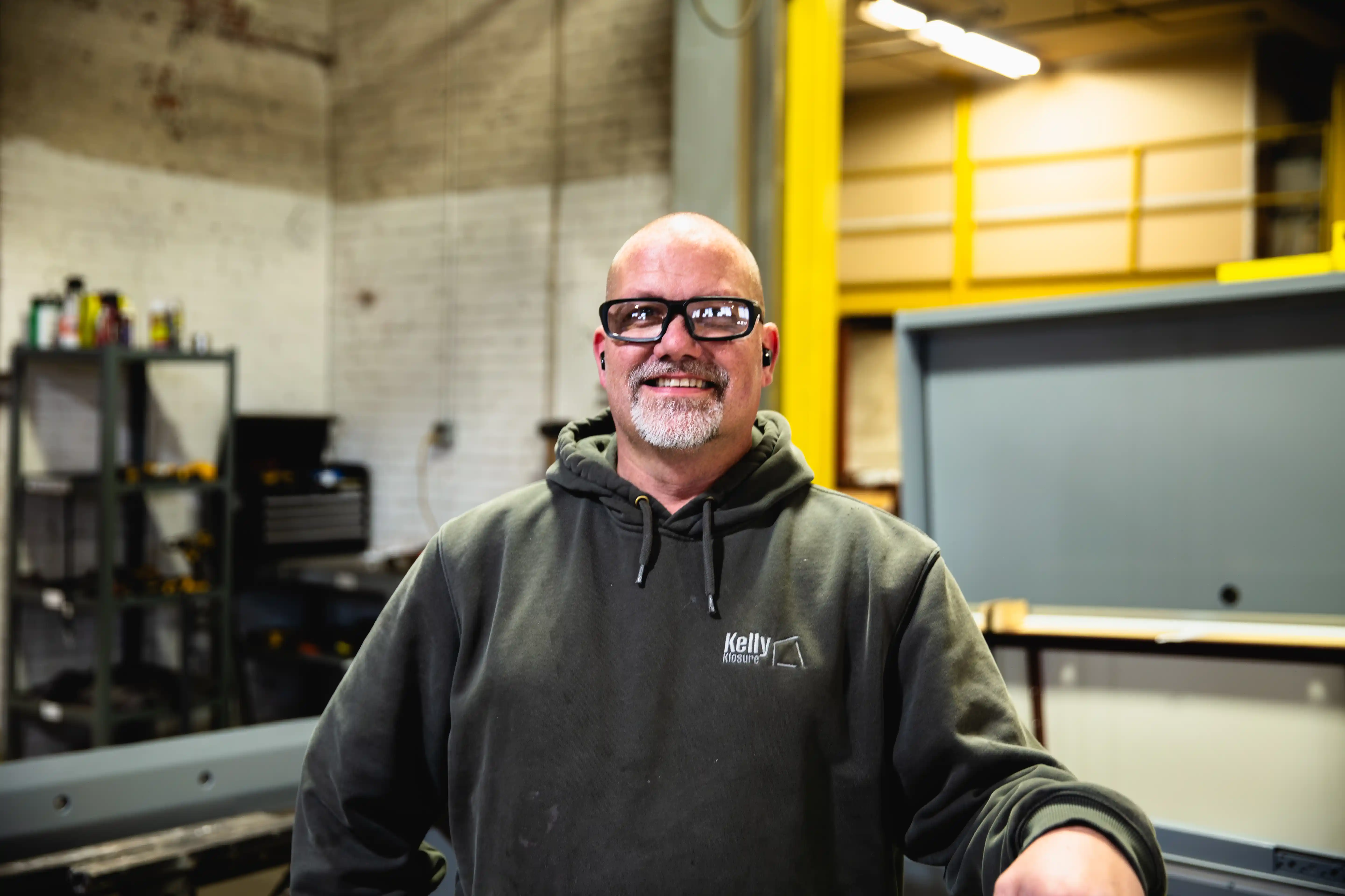 Smiling man wearing glasses and a Kelly Klosure hoodie in an industrial workshop.