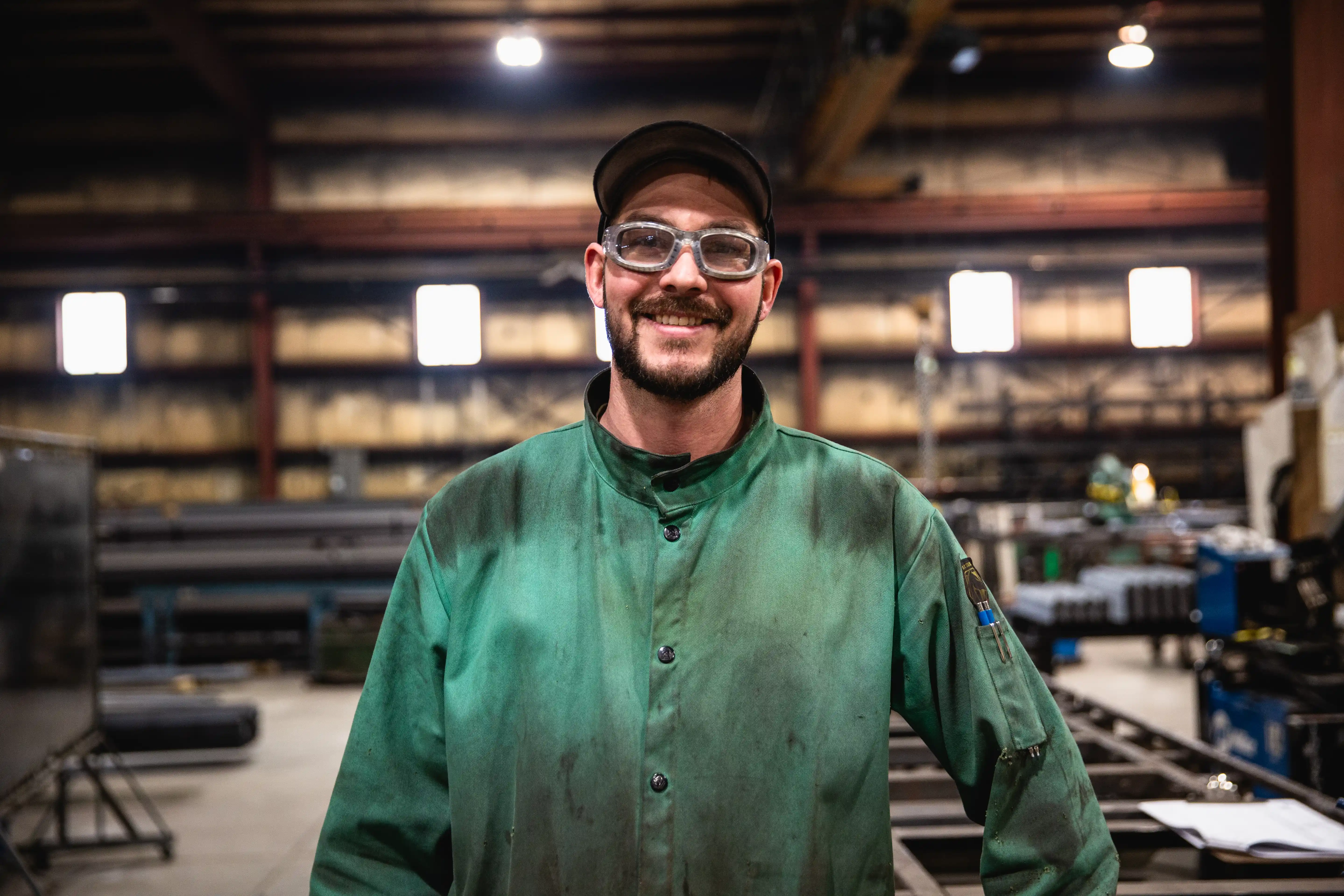 Smiling factory worker in safety glasses and a green work coat in an industrial workshop.