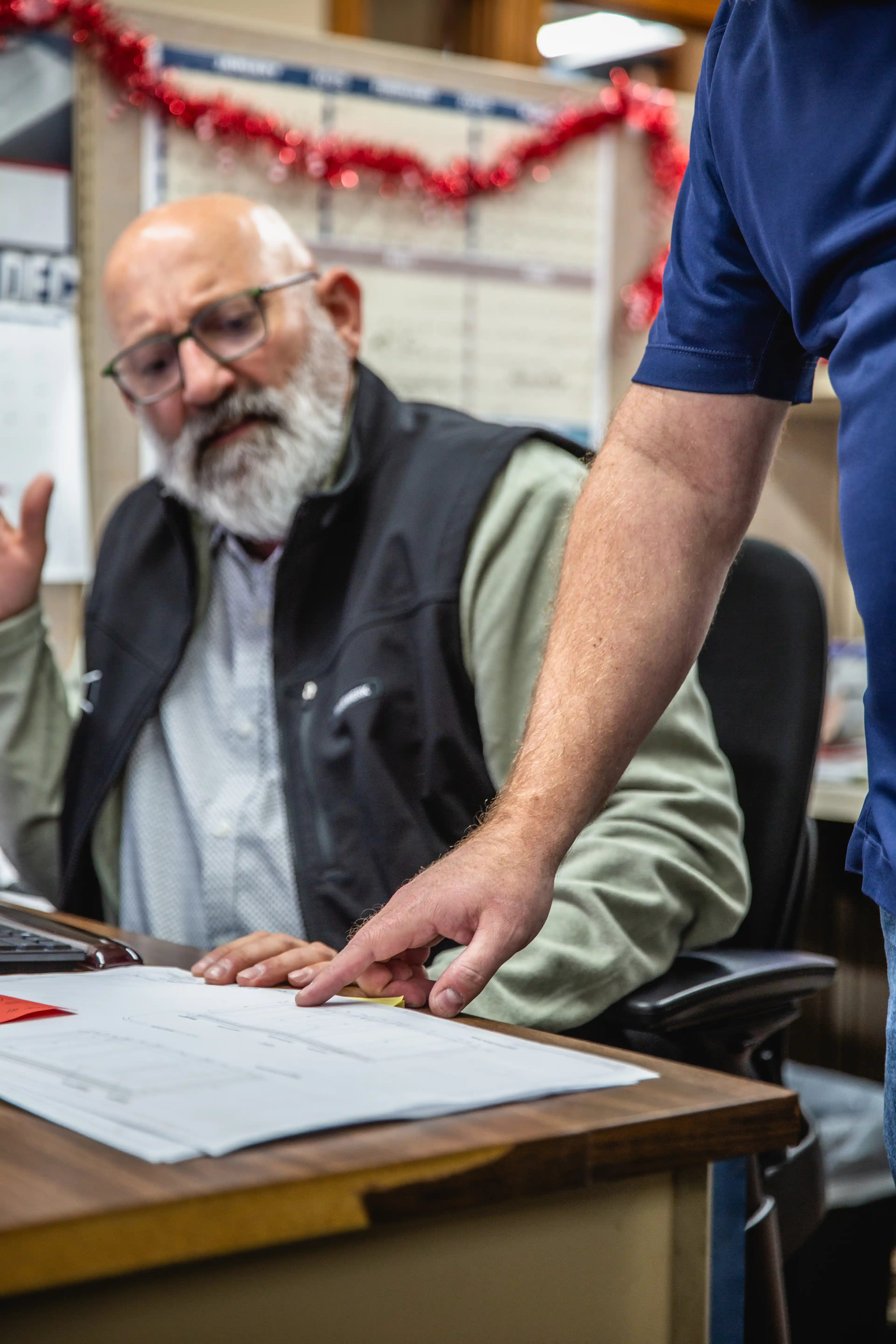Man in black vest gestures while another points at papers on a desk during discussion.