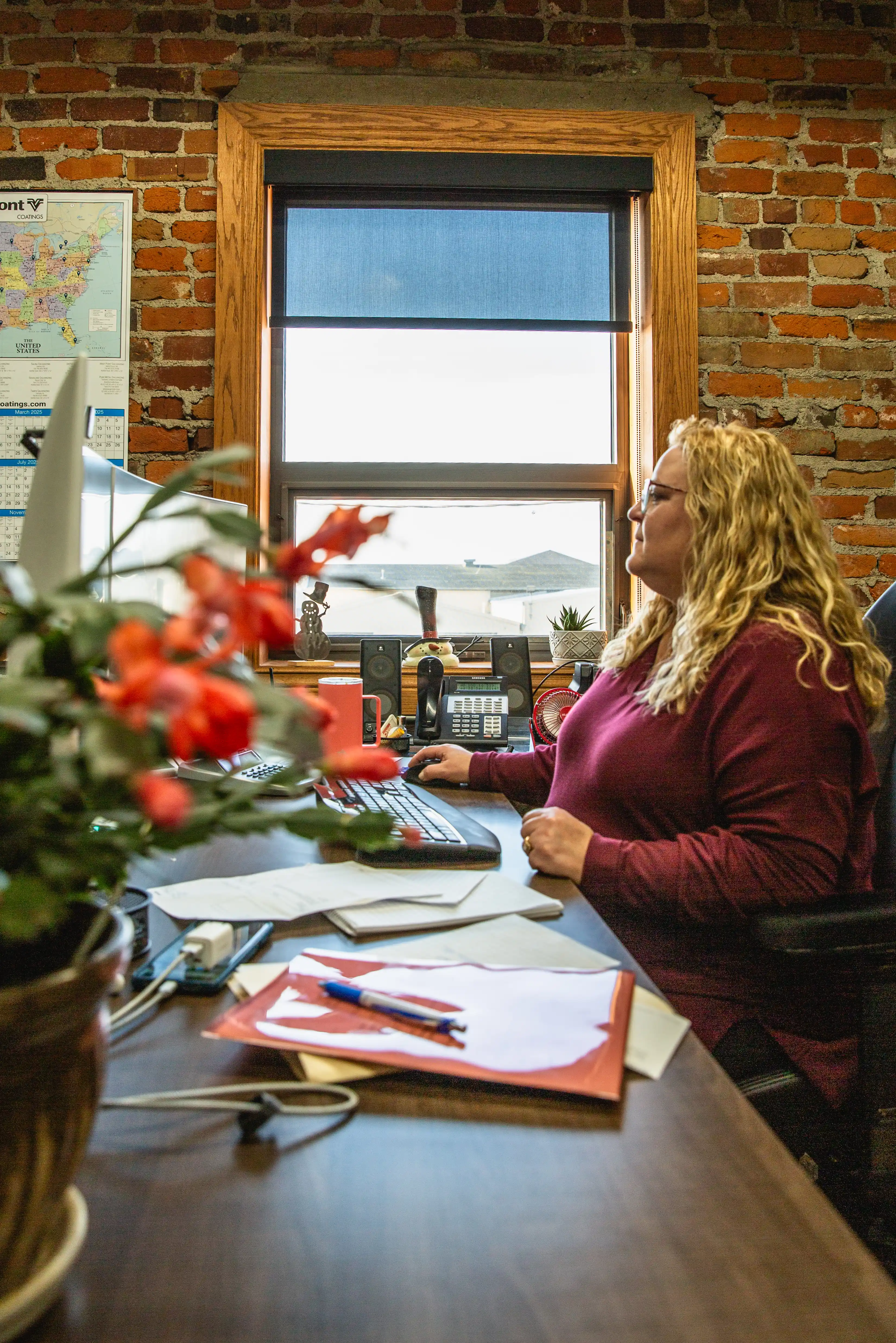 Woman with curly blond hair working at a desk with computer in an office with brick walls.