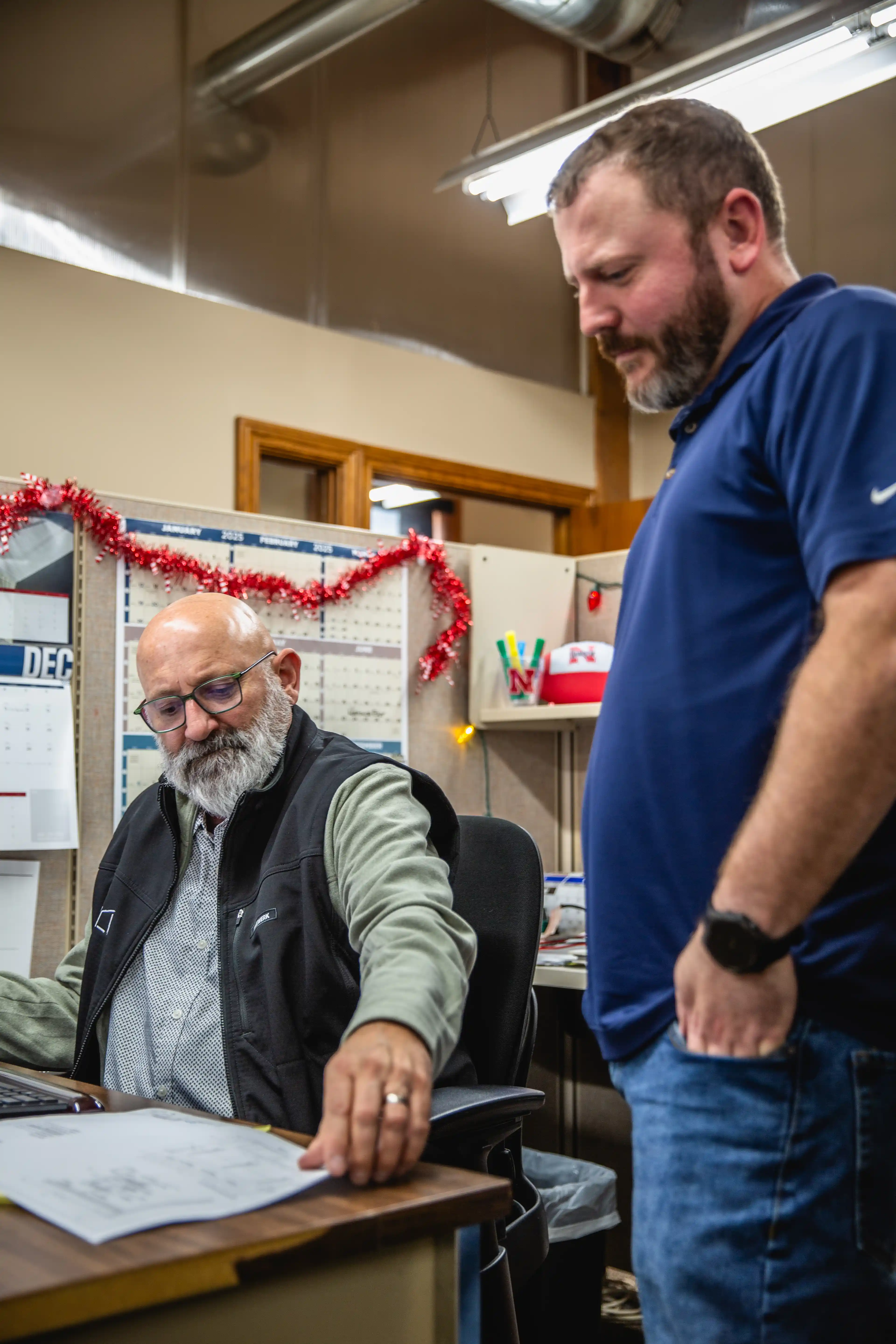 Two men in an office discuss papers, one seated with glasses and beard, the other standing.