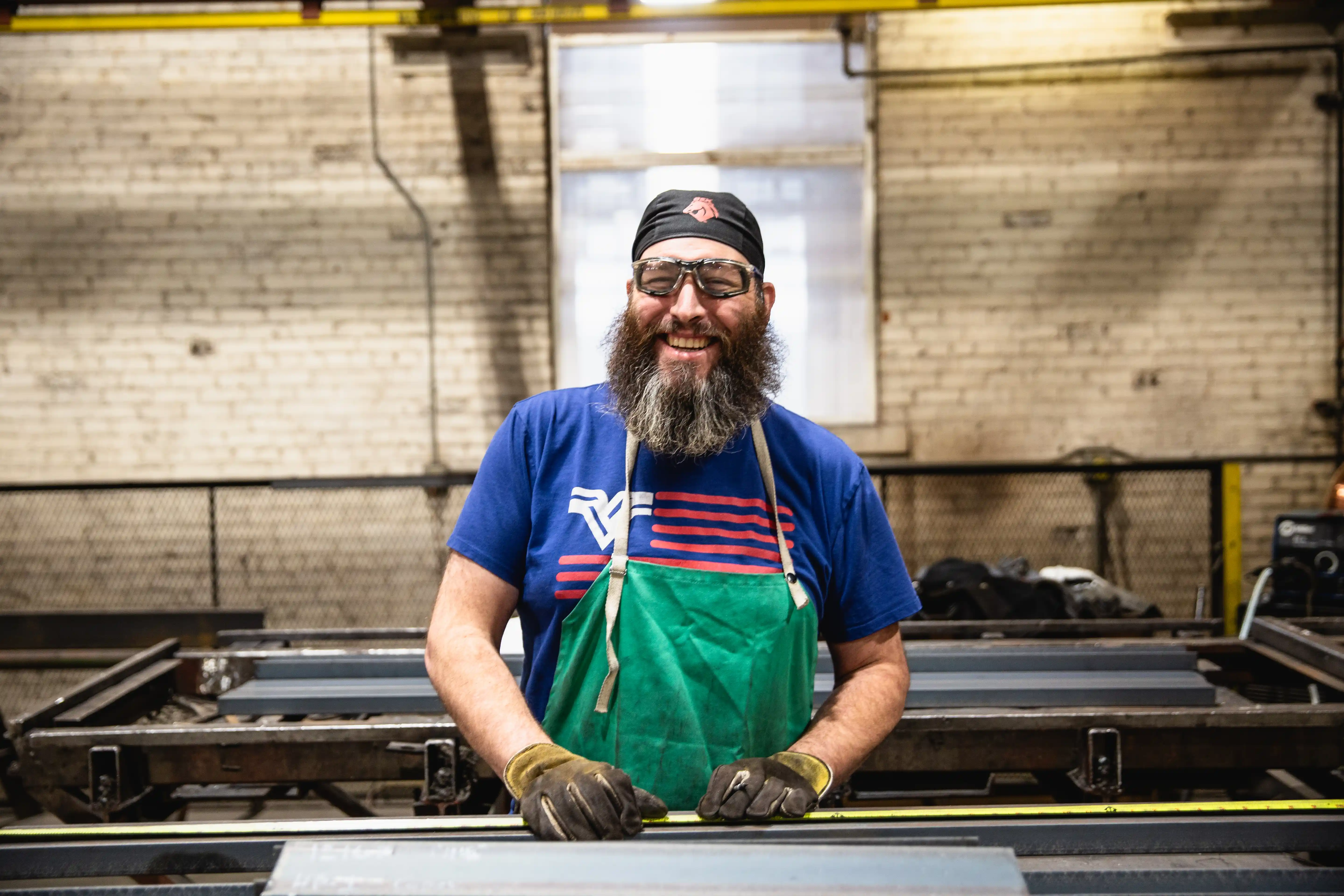 Smiling bearded man in safety glasses and apron measuring metal in an industrial workshop.