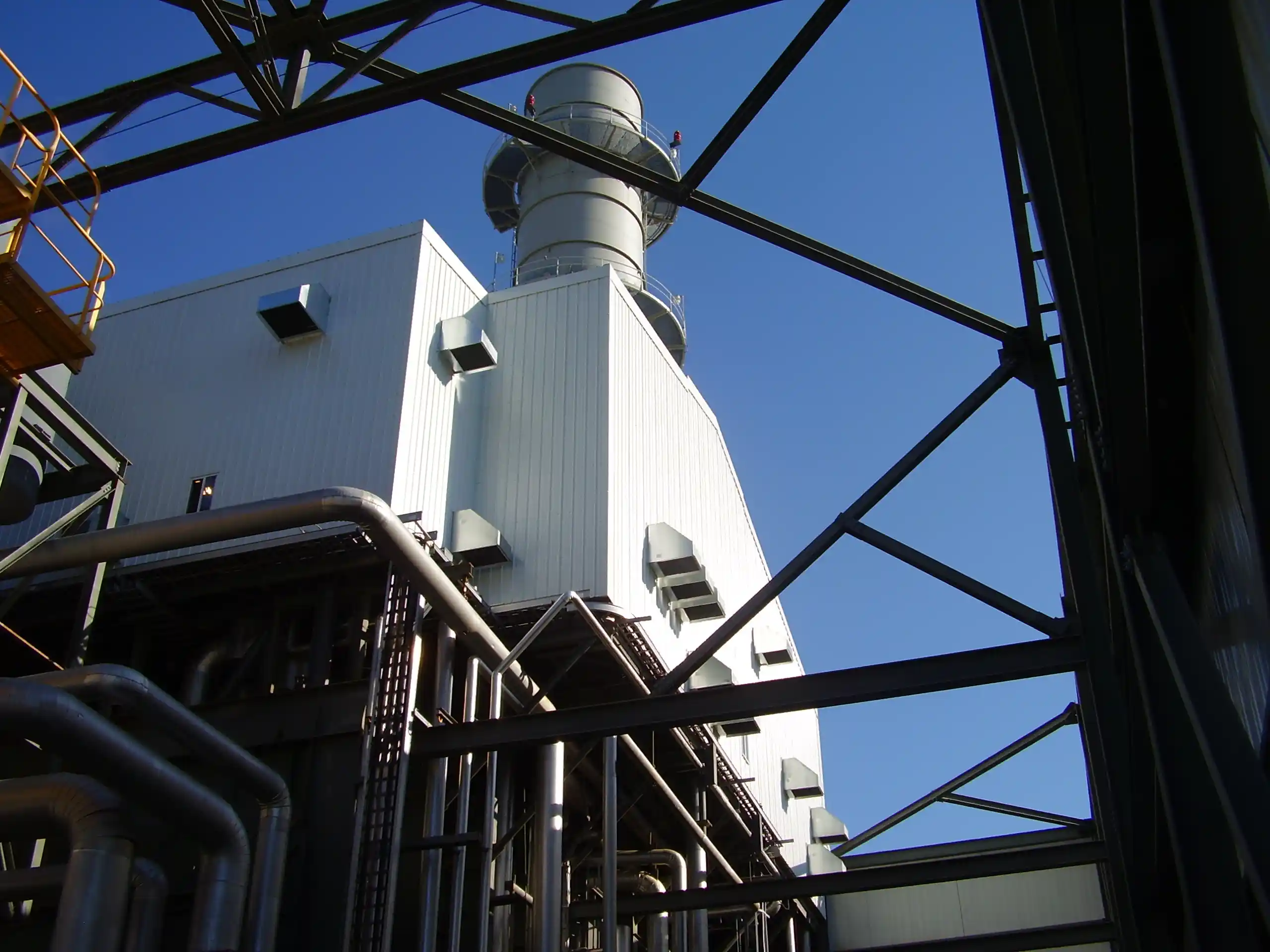 Industrial building with a large smokestack, metal pipes, and support beams under a clear blue sky.