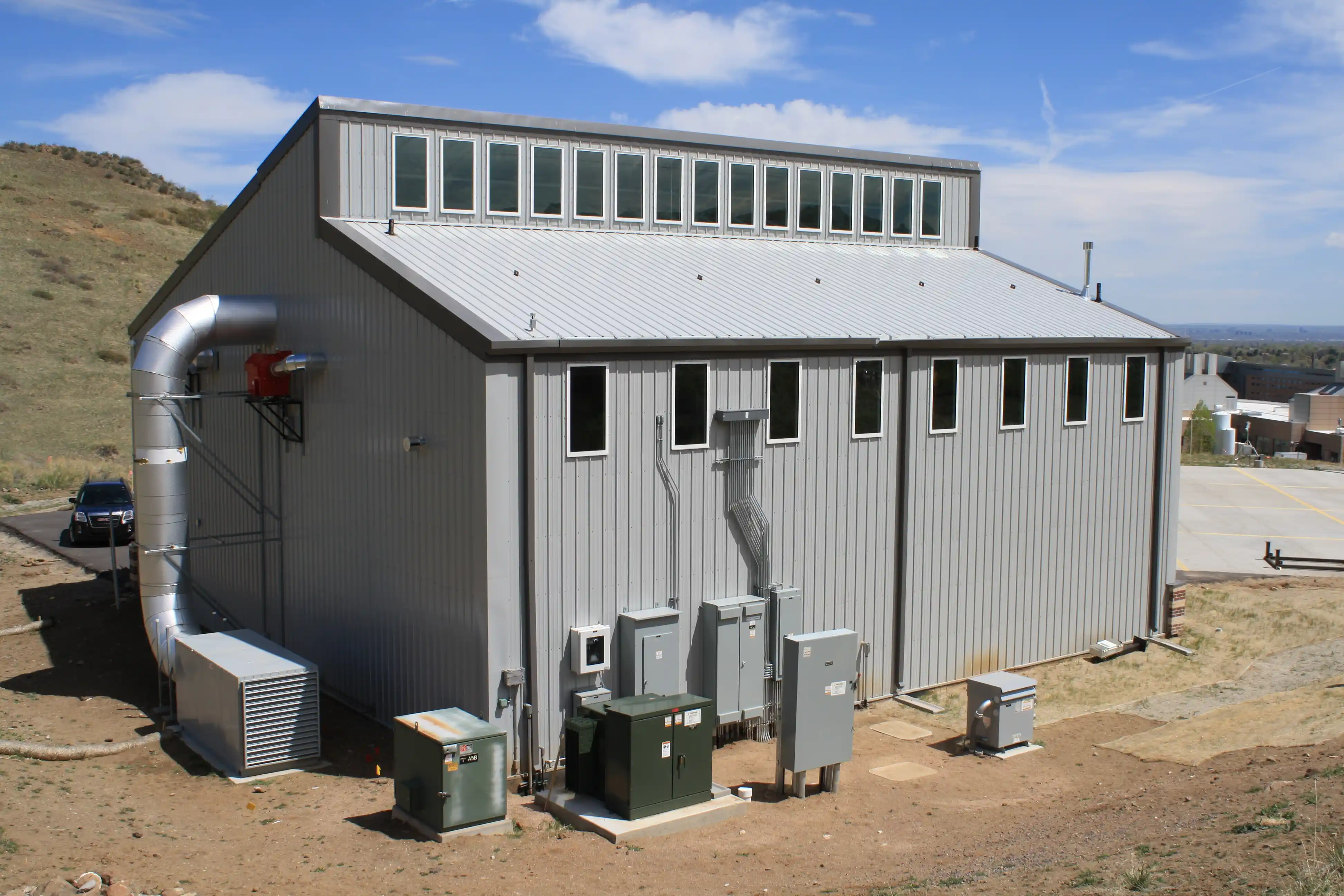 Metal industrial building with multiple small windows, ventilation pipe, and electrical equipment outside.