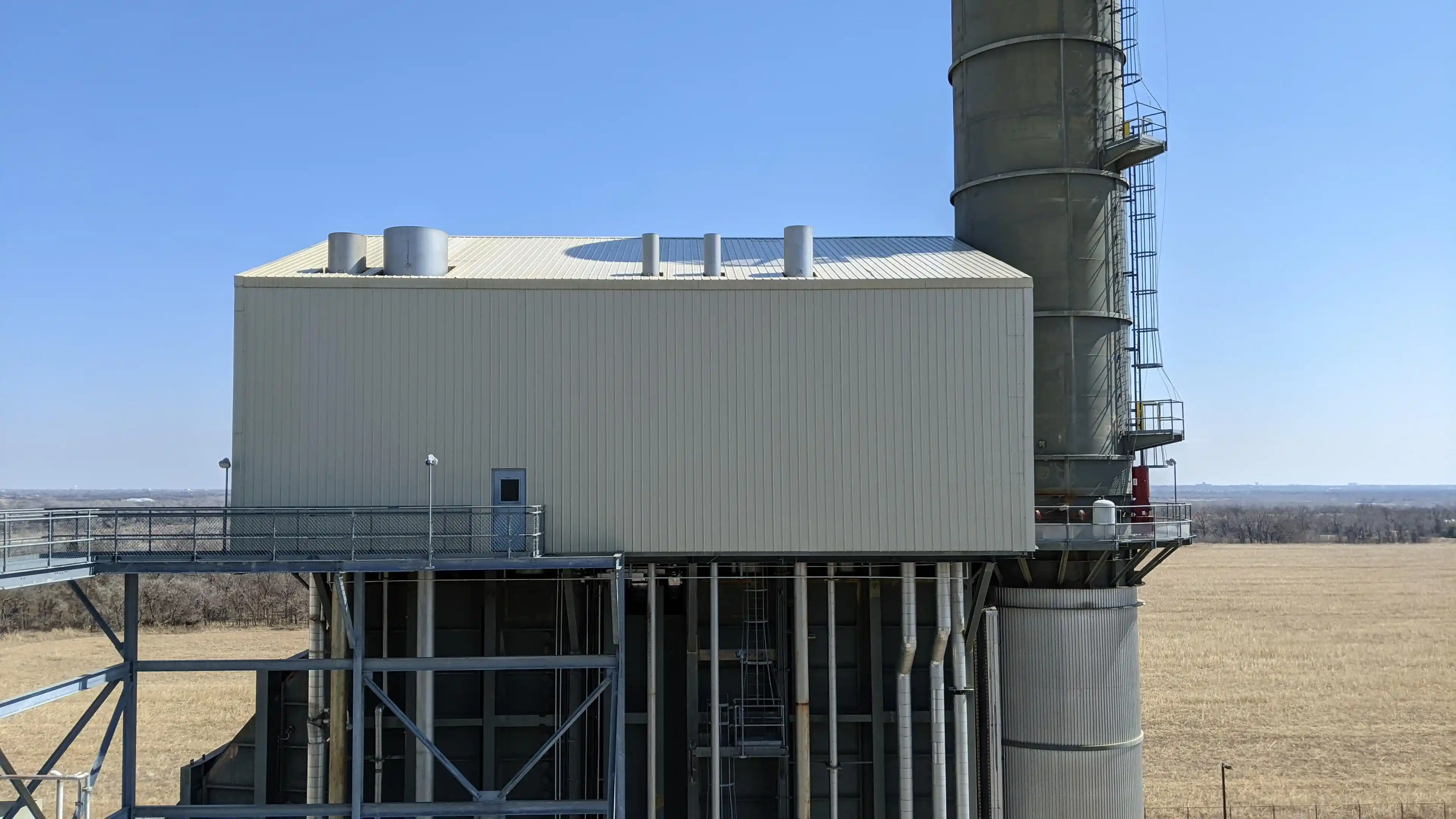 Industrial building with pipes and a tall cylindrical structure in a rural open field under clear sky.