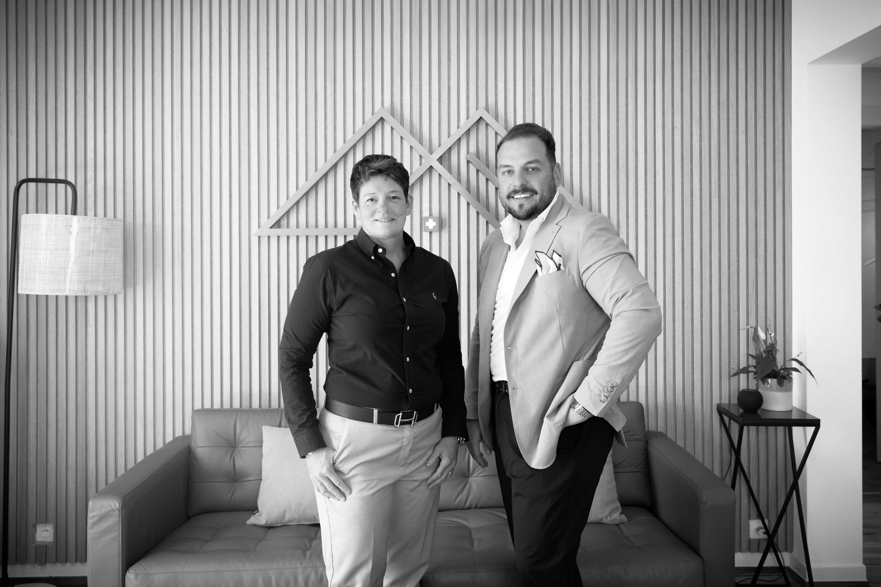 Deux personnes souriantes dans un bureau avec une table, chaises et photos de voiture de course et logo immobilier.