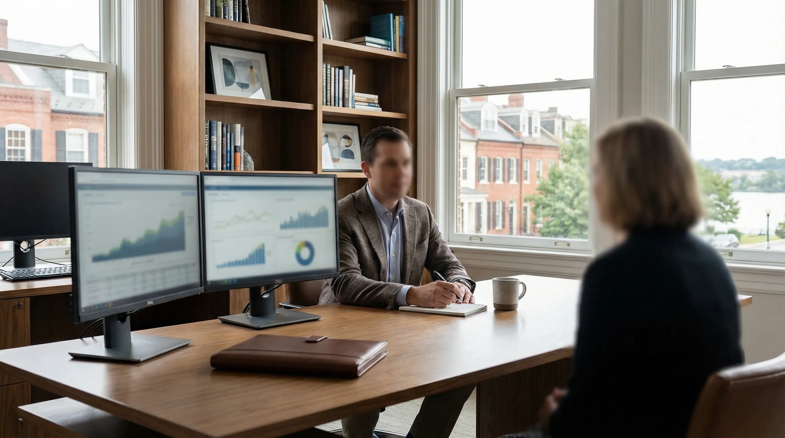 Professional financial advisor in a well-appointed consultation office in Alexandria, VA meeting with a client at a polished desk with portfolio documents visible