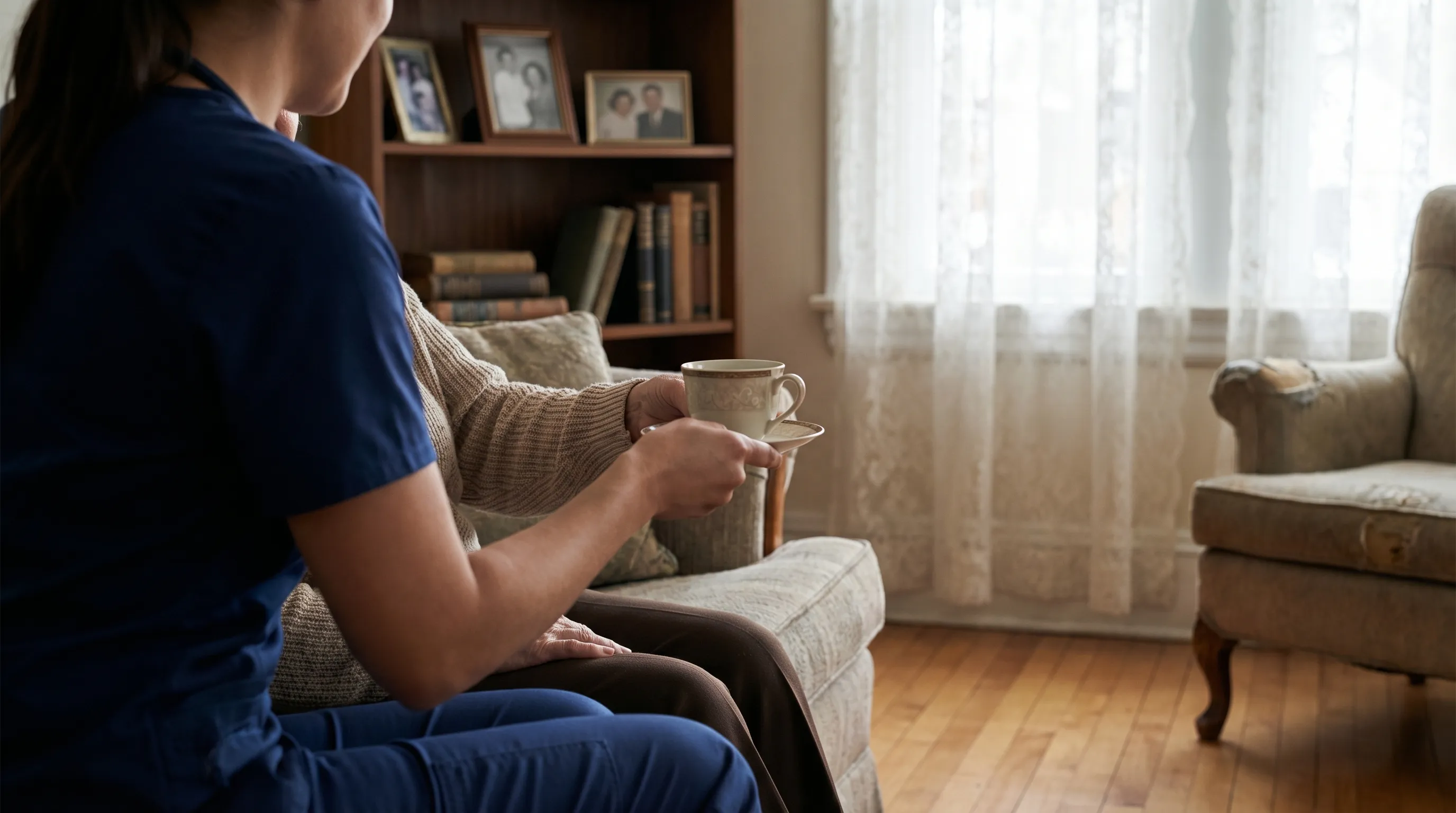 Warm afternoon in a Waterbury CT living room, caregiver in professional scrubs helping an elderly woman with tea, older home setting with family photos and natural light signaling dignity and care