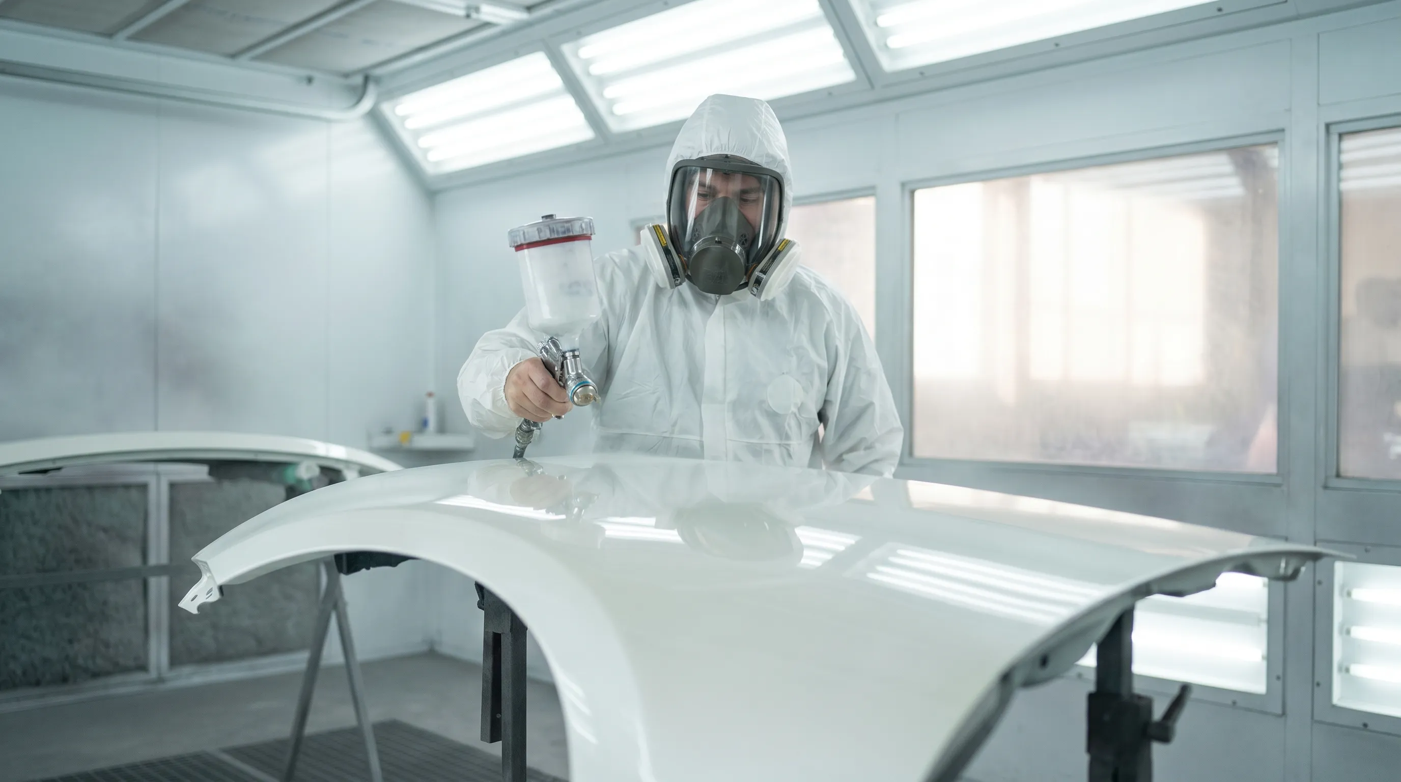 Auto body technician in protective spray gear applying paint to a vehicle panel inside a modern Erie, PA collision shop