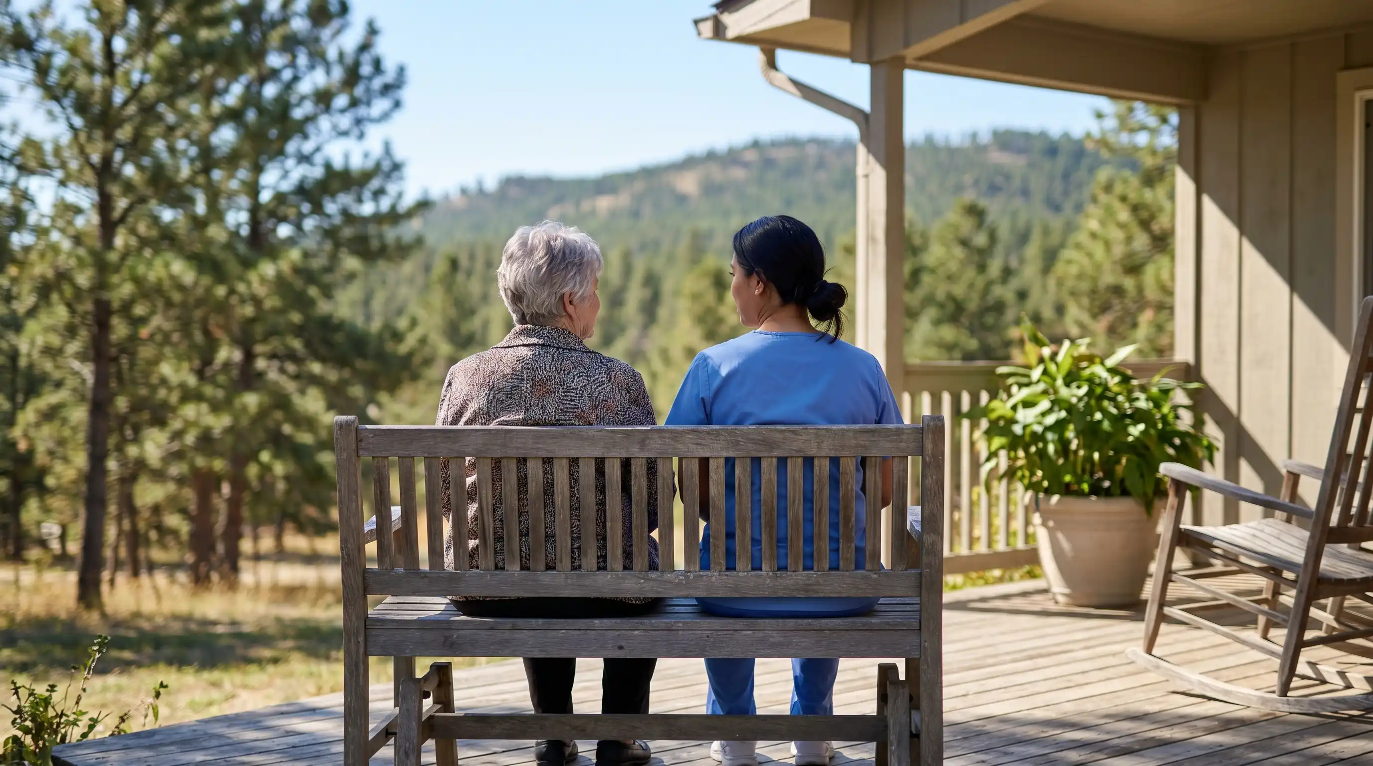Professional senior home care aide in conversation with an elderly woman on the porch of a ranch-style home in Rapid City, SD, with Black Hills ponderosa pines in the background