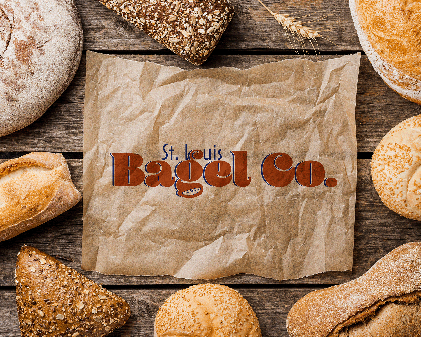 Logo for St. Louis Bagel Co. rendered on a crumpled and unfolded piece of parchment paper and surrounded by different types of bread with a flat wooden background.