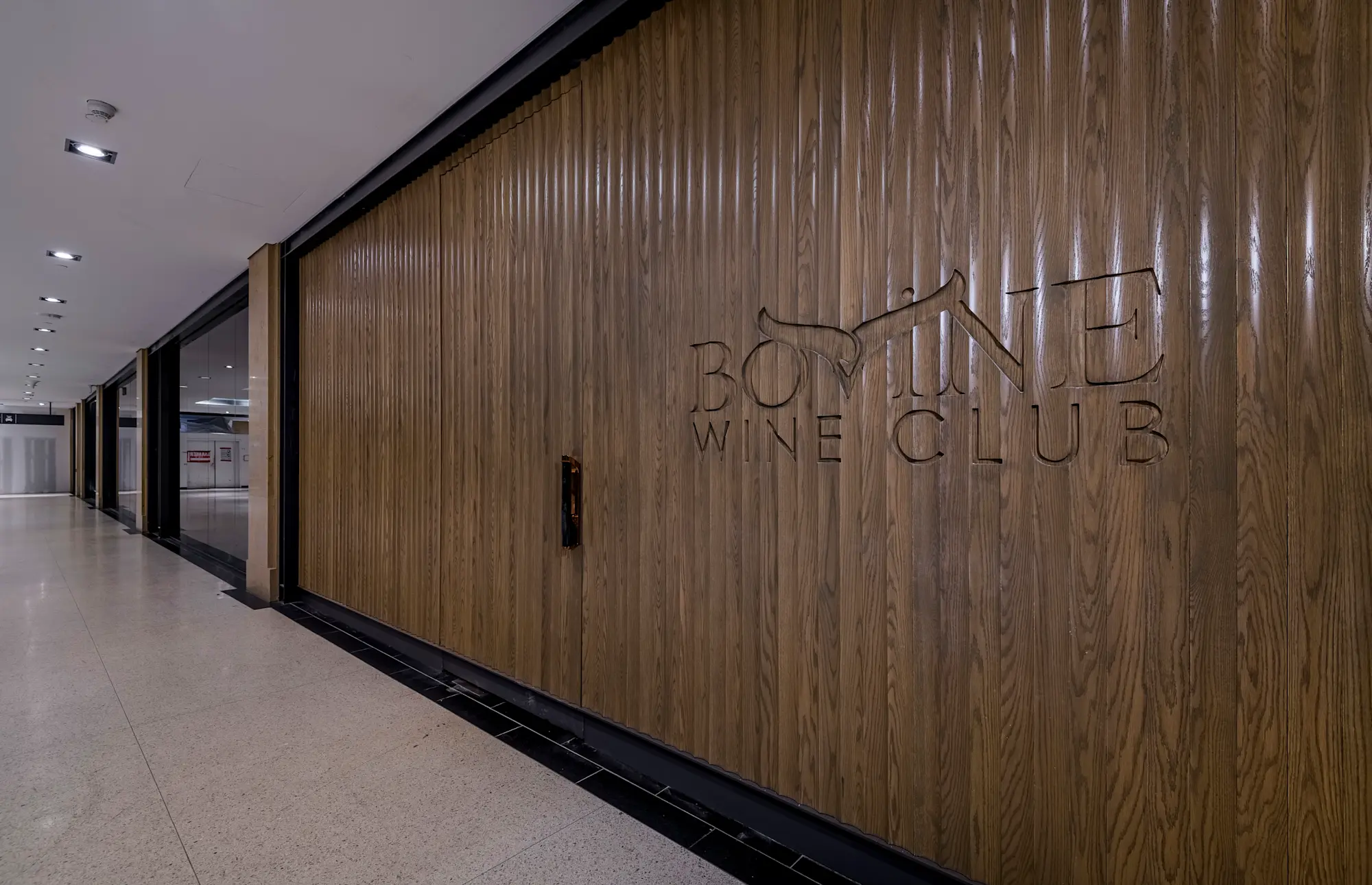 Wood-paneled wall with carved text reading 'Bovine Wine Club' in a modern indoor hallway.