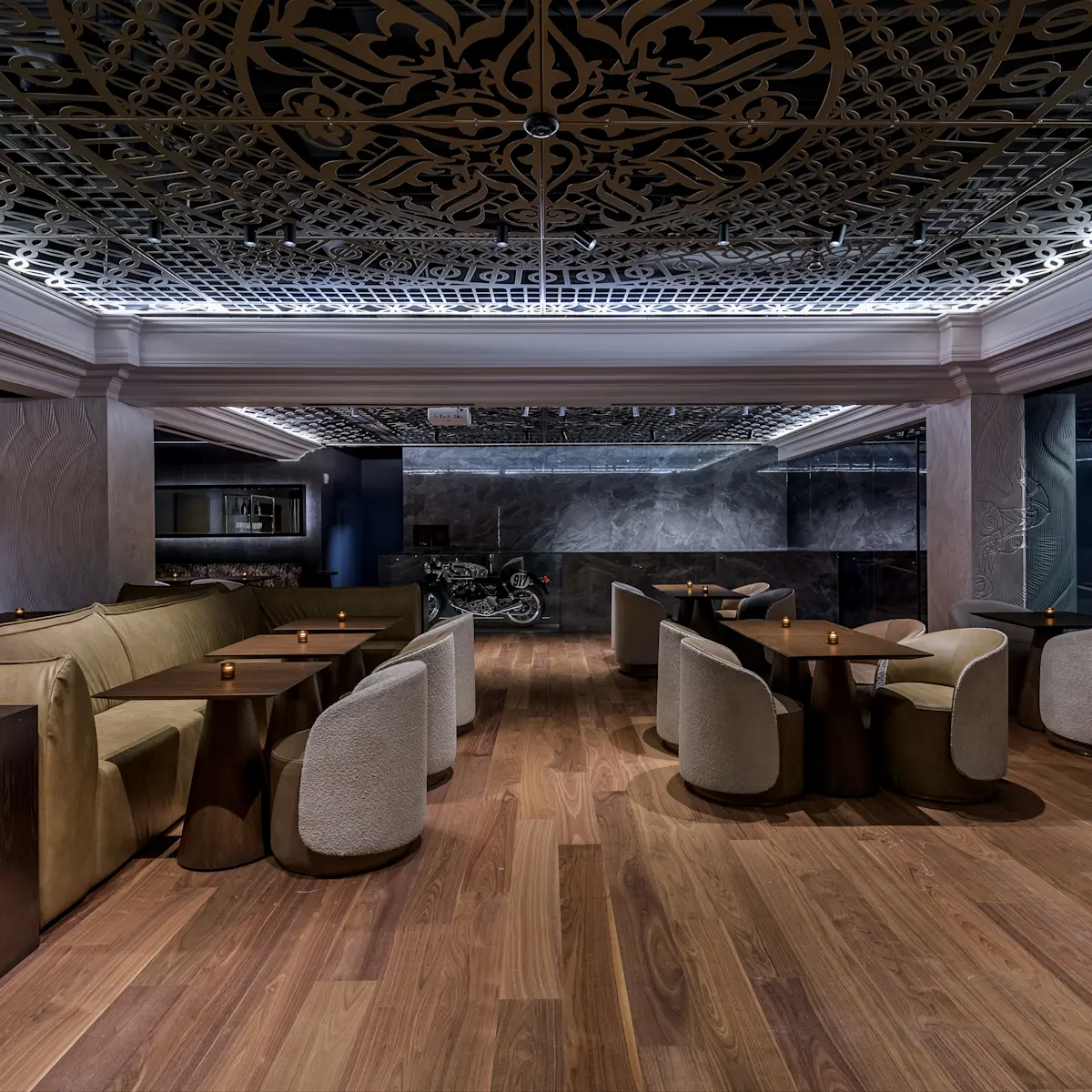 Modern lounge area with wooden floors, cushioned chairs, long leather sofas, dark wooden tables, intricate patterned ceiling, and a motorcycle displayed against a marble wall.