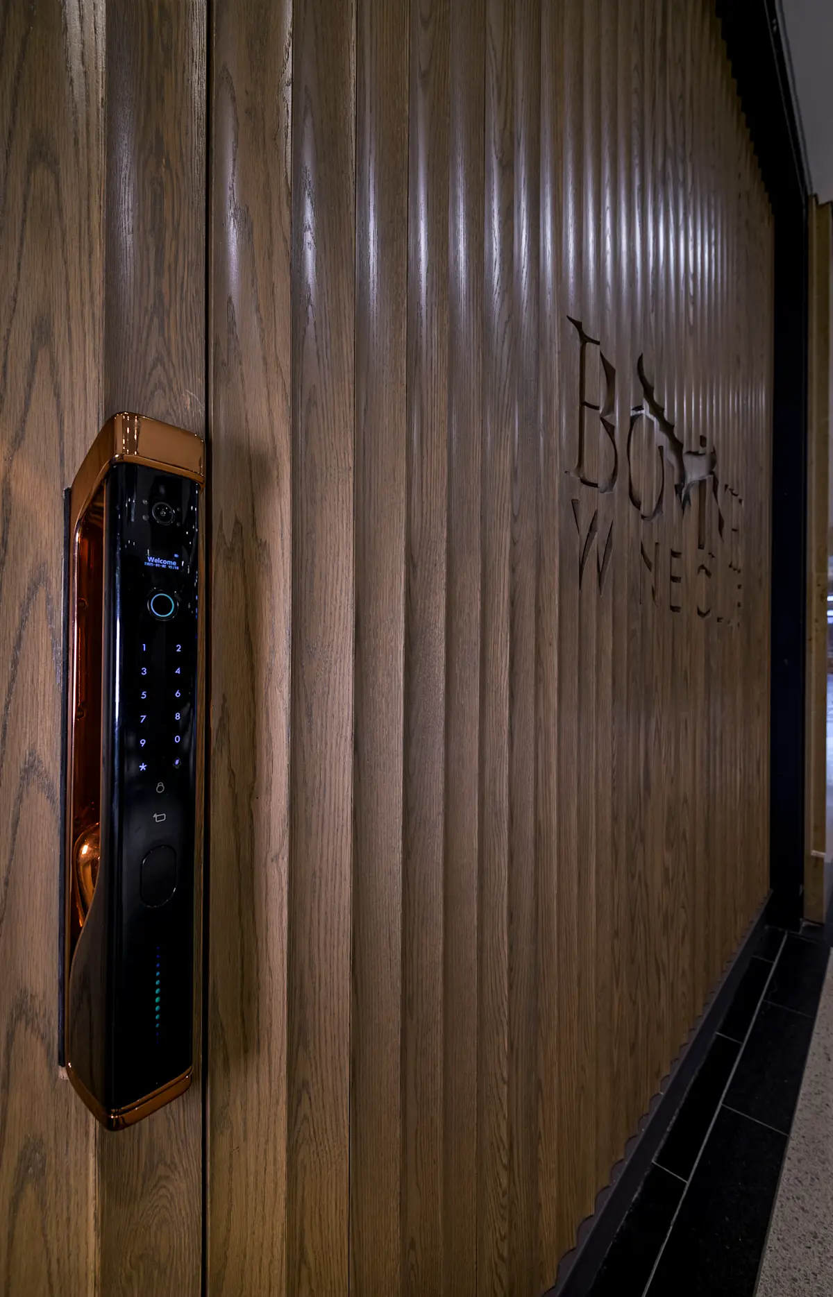 Close-up of a modern electronic door lock on a vertically slatted wooden door with 'Bovine Wines' carved into the wood.