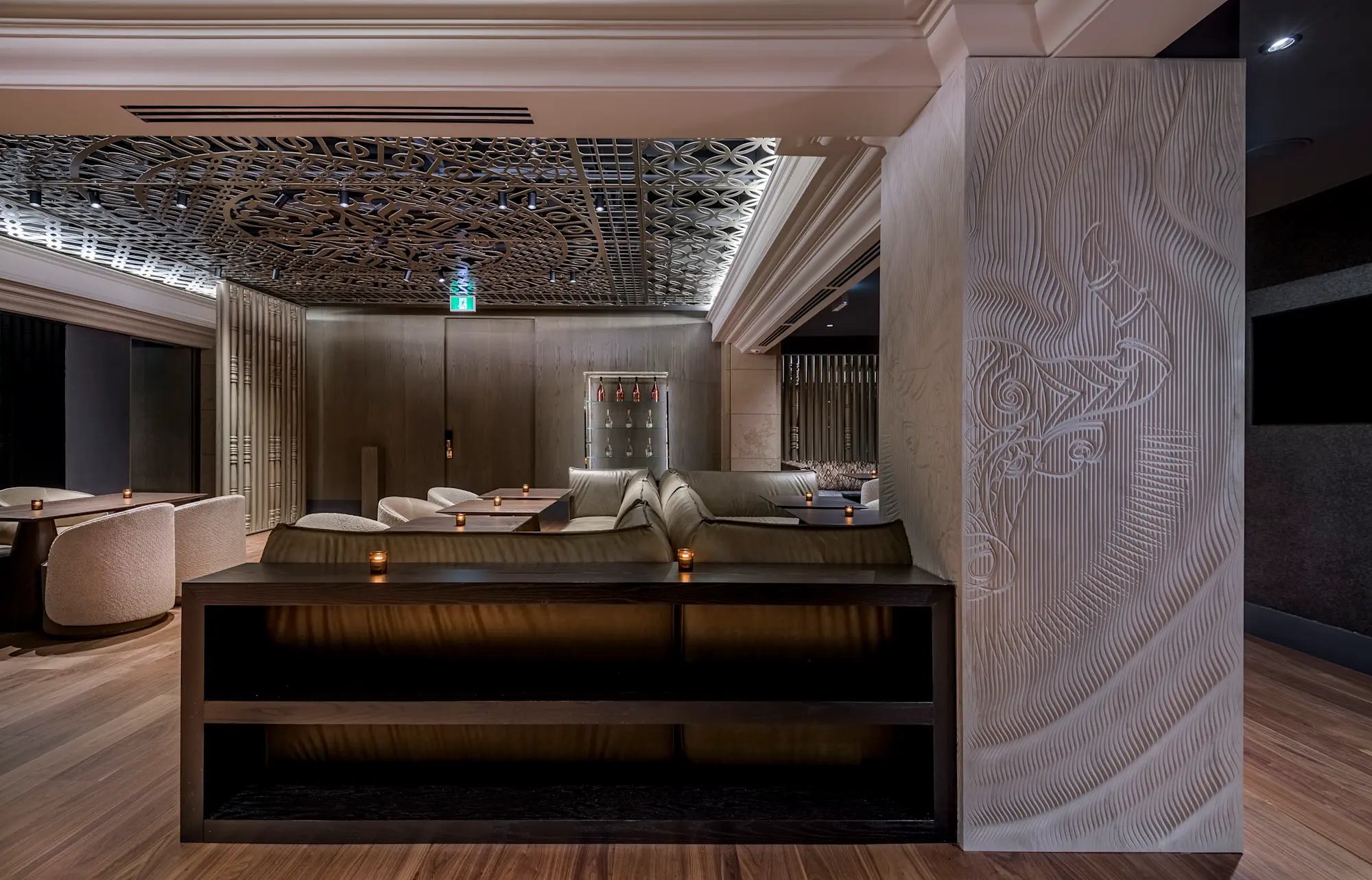 Elegant lounge area with beige cushioned seats, candlelit tables, wooden flooring, and a ceiling featuring intricate circular metal patterns.