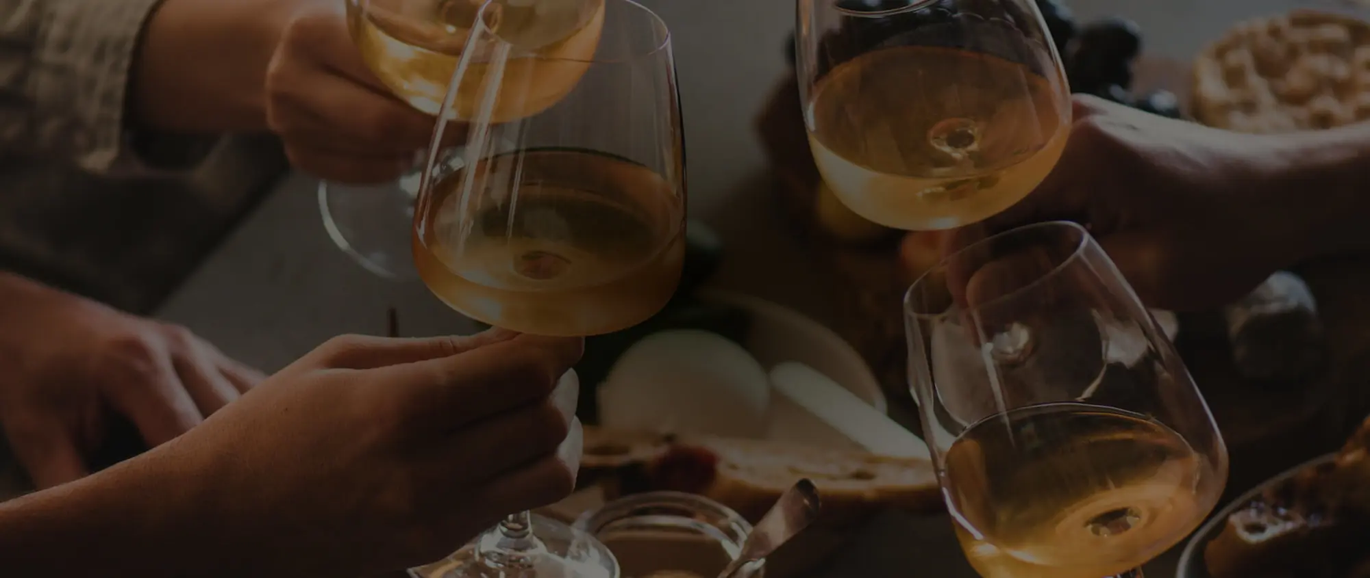 Four hands clinking glasses of white wine over a table with bread and snacks.