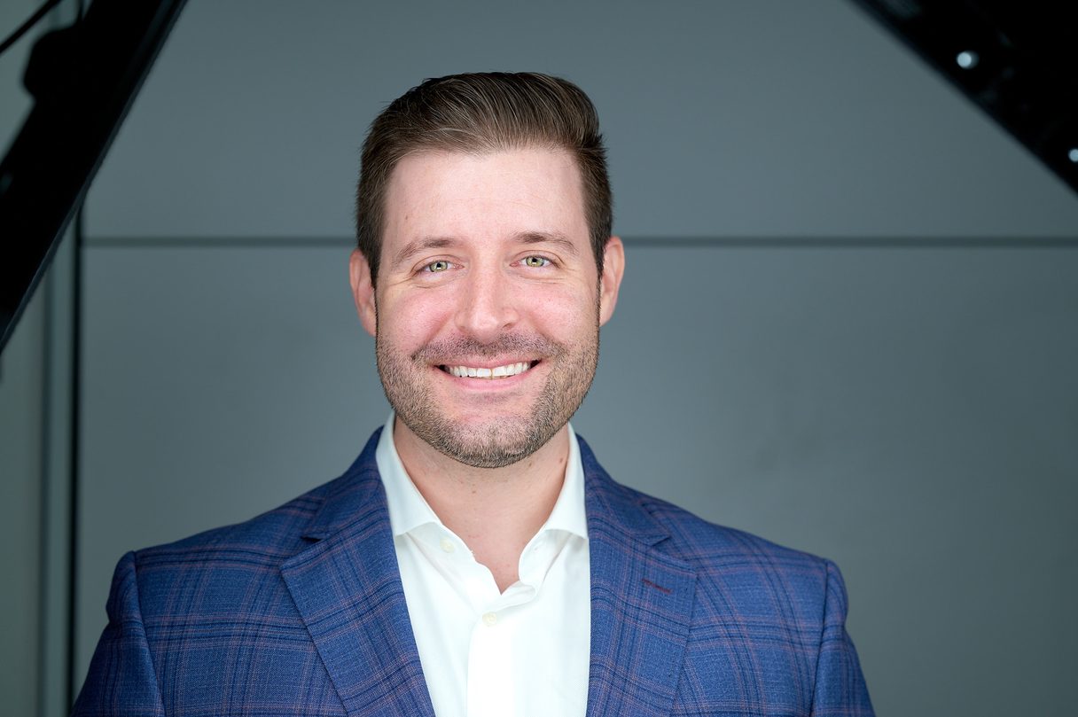 Smiling man with short brown hair and beard wearing a blue checkered blazer and white shirt against a gray background.