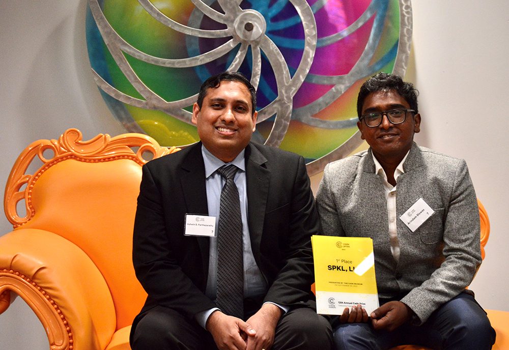 Two men in suits sitting on an orange ornate couch, one holding a yellow award plaque for 1st place in the 12th Annual Code Prize.