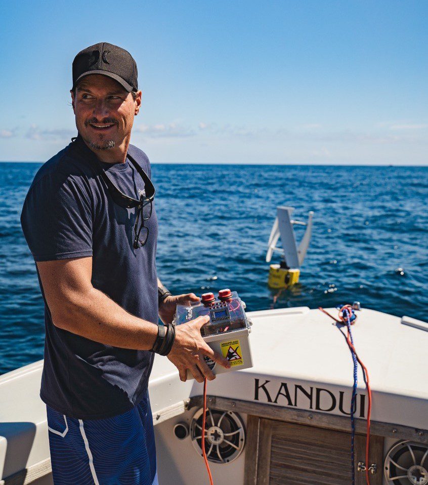 Man in navy shirt and cap holding a small electronic device while standing on a boat named KANDUI in the ocean.
