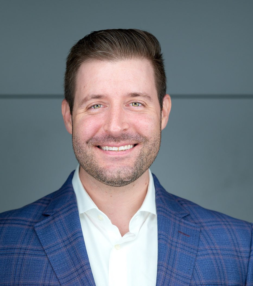 Smiling man with short brown hair and beard wearing a blue checkered suit jacket and white shirt against a gray background.
