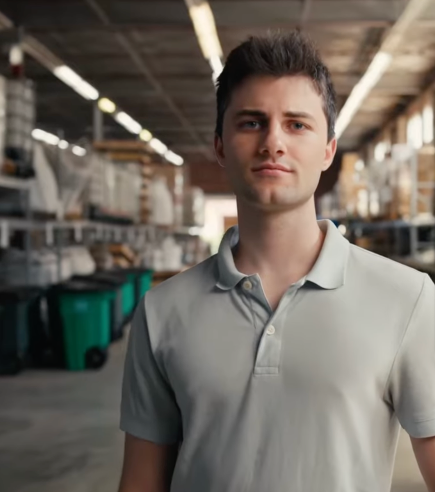 Young man with dark hair wearing a light gray polo shirt standing inside a warehouse with shelves and green bins.