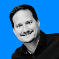 Smiling man with short dark hair and goatee wearing a black shirt against a bright blue background.