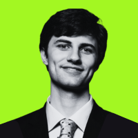 Young man with dark hair wearing a black suit, light blue shirt, and patterned tie, smiling against a green background.