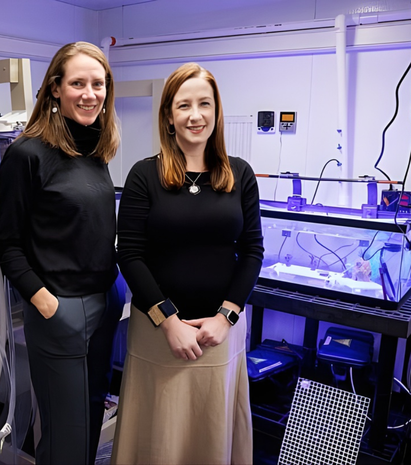 Two women smiling and standing in a lab with illuminated scientific equipment and tanks in the background.