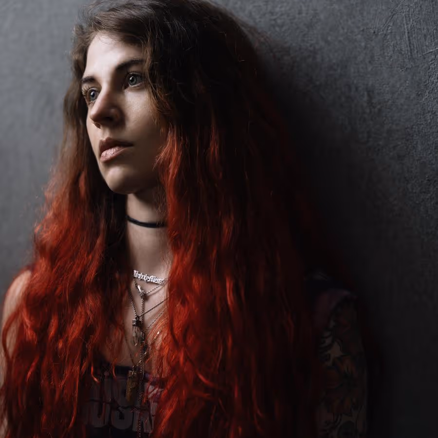 Portrait of a person, leaning against a wall and looking off into the distance. The person has long brown and red hair.