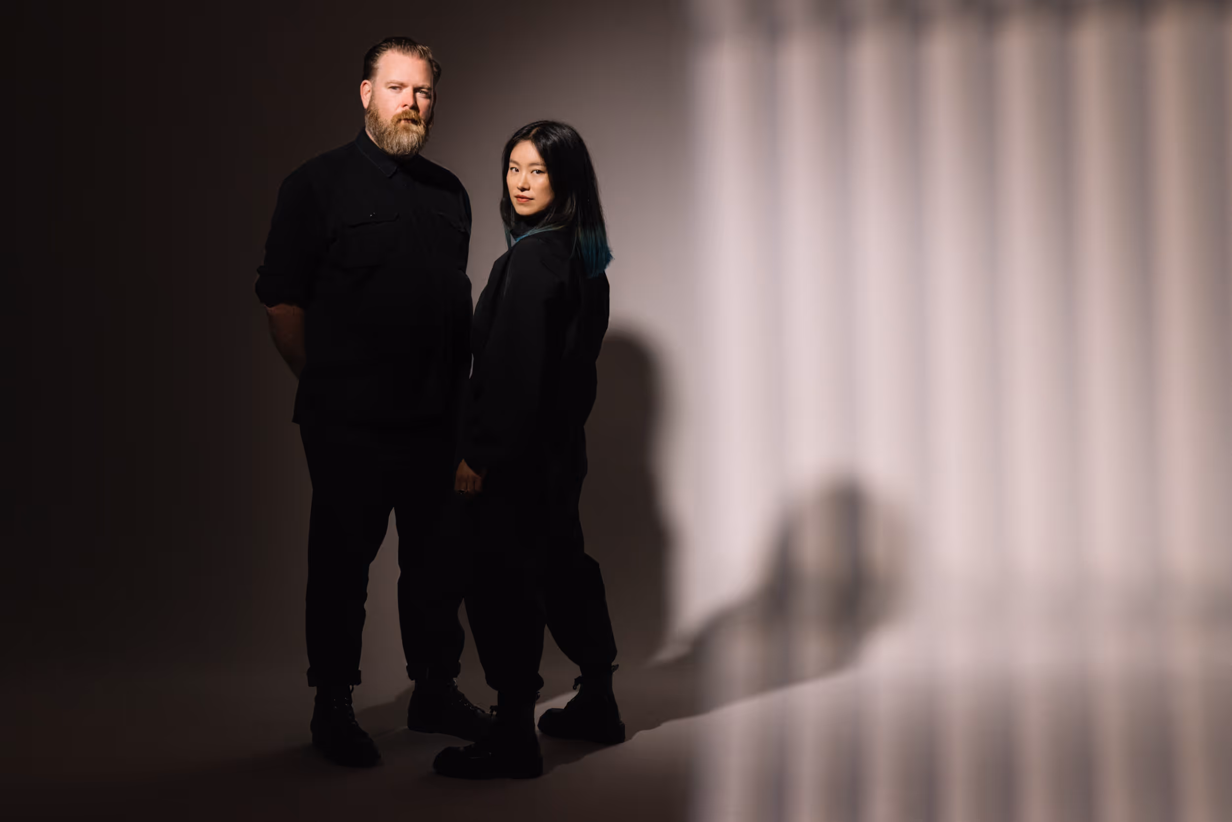 Two people standing, facing the camera. There is cinematic lighting hitting their faces, and creating vertical light shadows on the wall. 