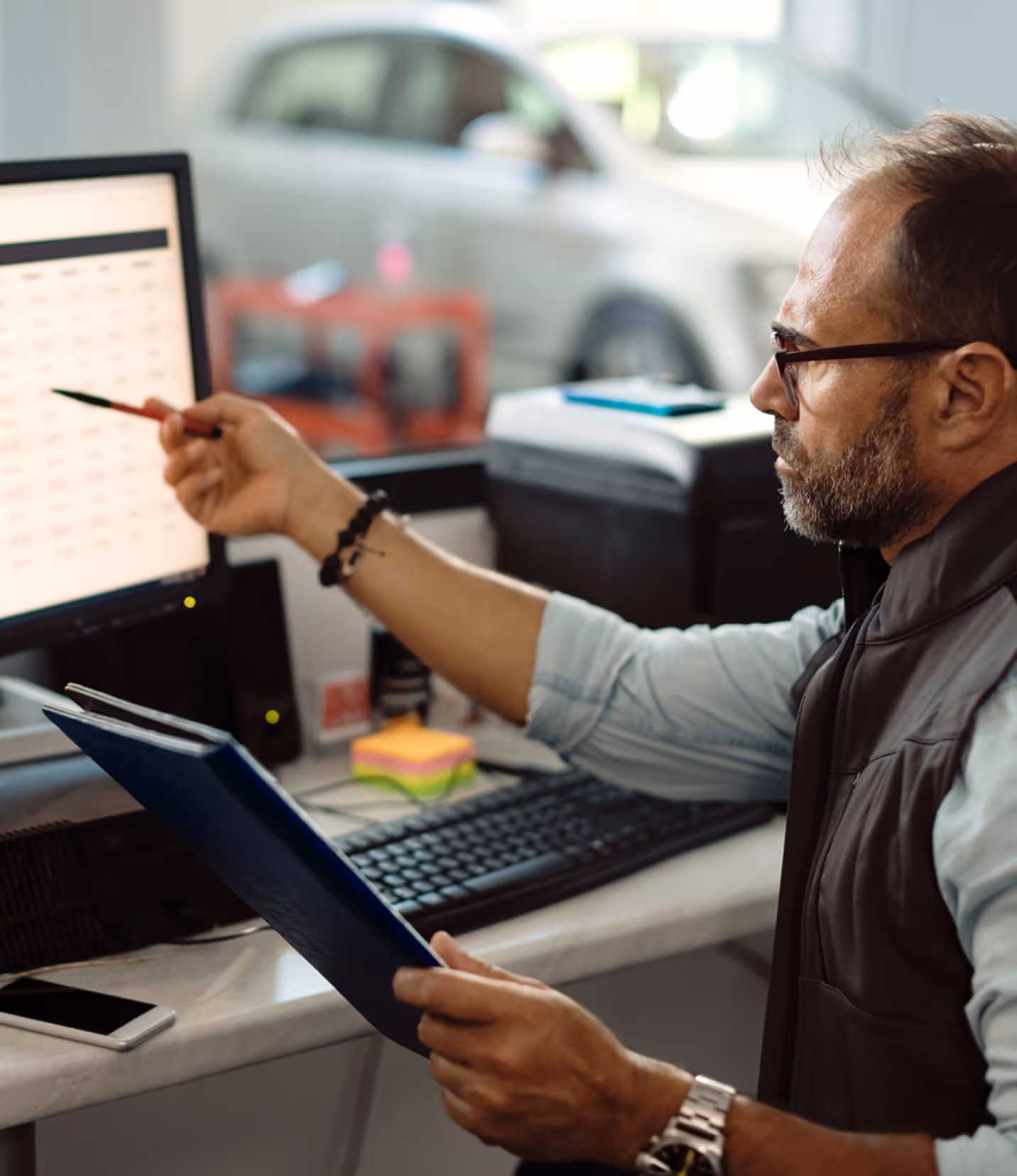 Man in glasses pointing at computer screen while holding a folder in an office setting.