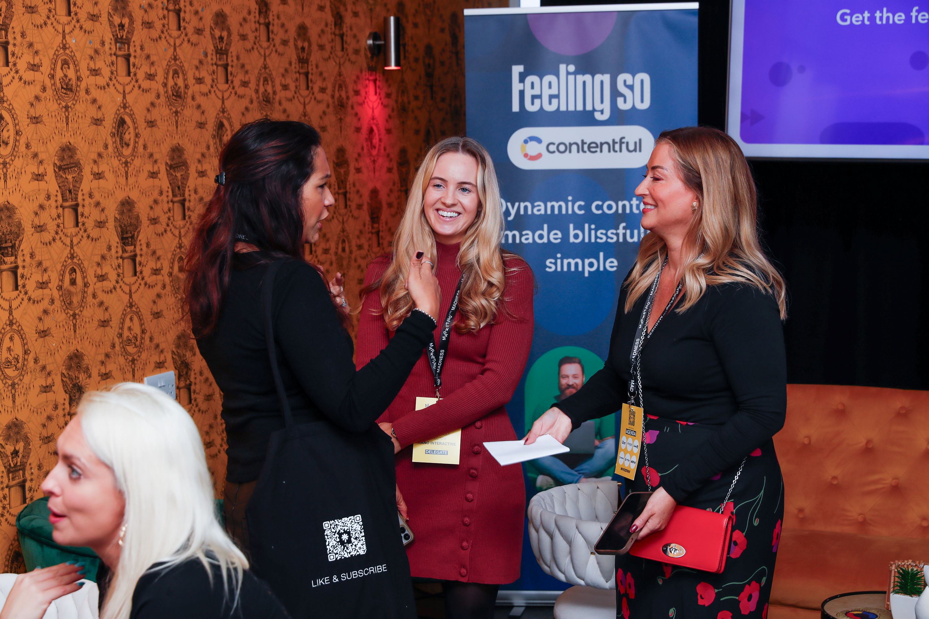 A group of women stand together chatting and laughing at an event, with warm patterned wallpaper behind them and a branded Contentful banner in the background. One attendee holds a red bag, another carries a black tote with a QR code, and the atmosphere feels friendly and relaxed.