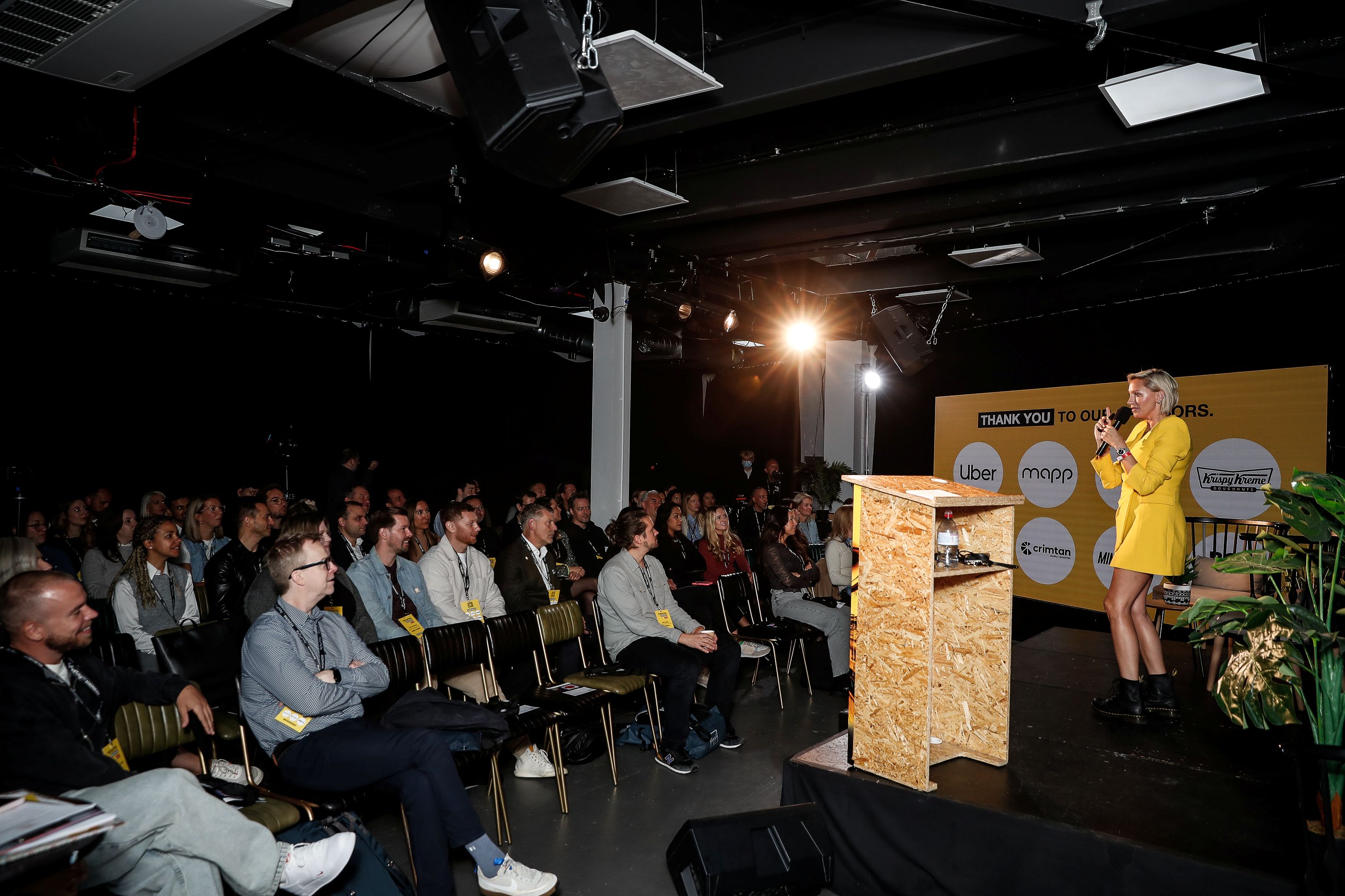 Katie Street wearing a bright yellow outfit presents on stage to a full seated audience in a dark event space. A yellow sponsor backdrop and a small wooden lectern are on stage.