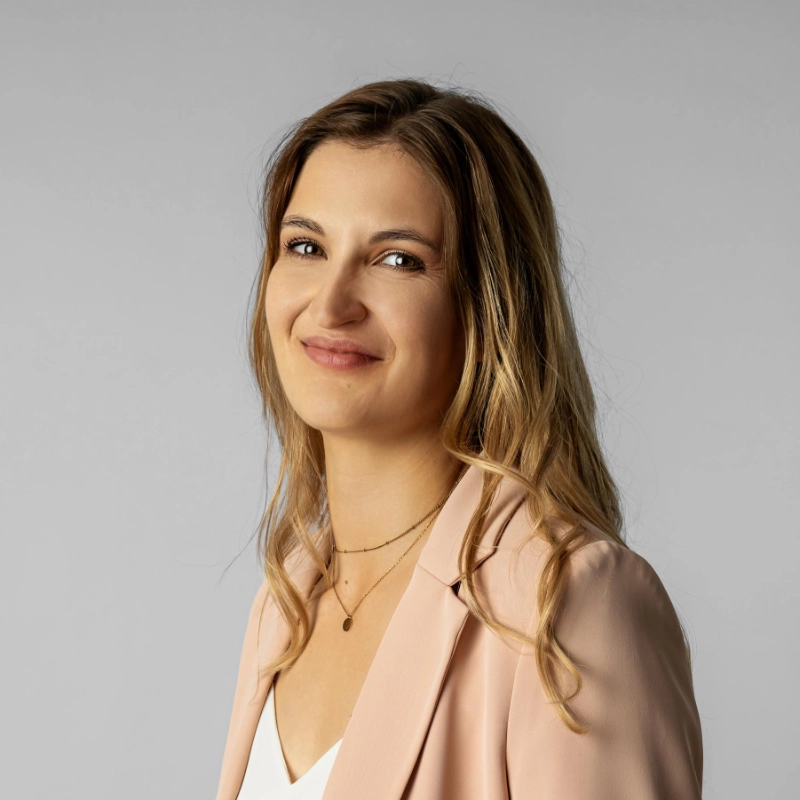 Porträt einer lächelnden Frau mit langen blonden Haaren, die einen hellrosa Blazer trägt.