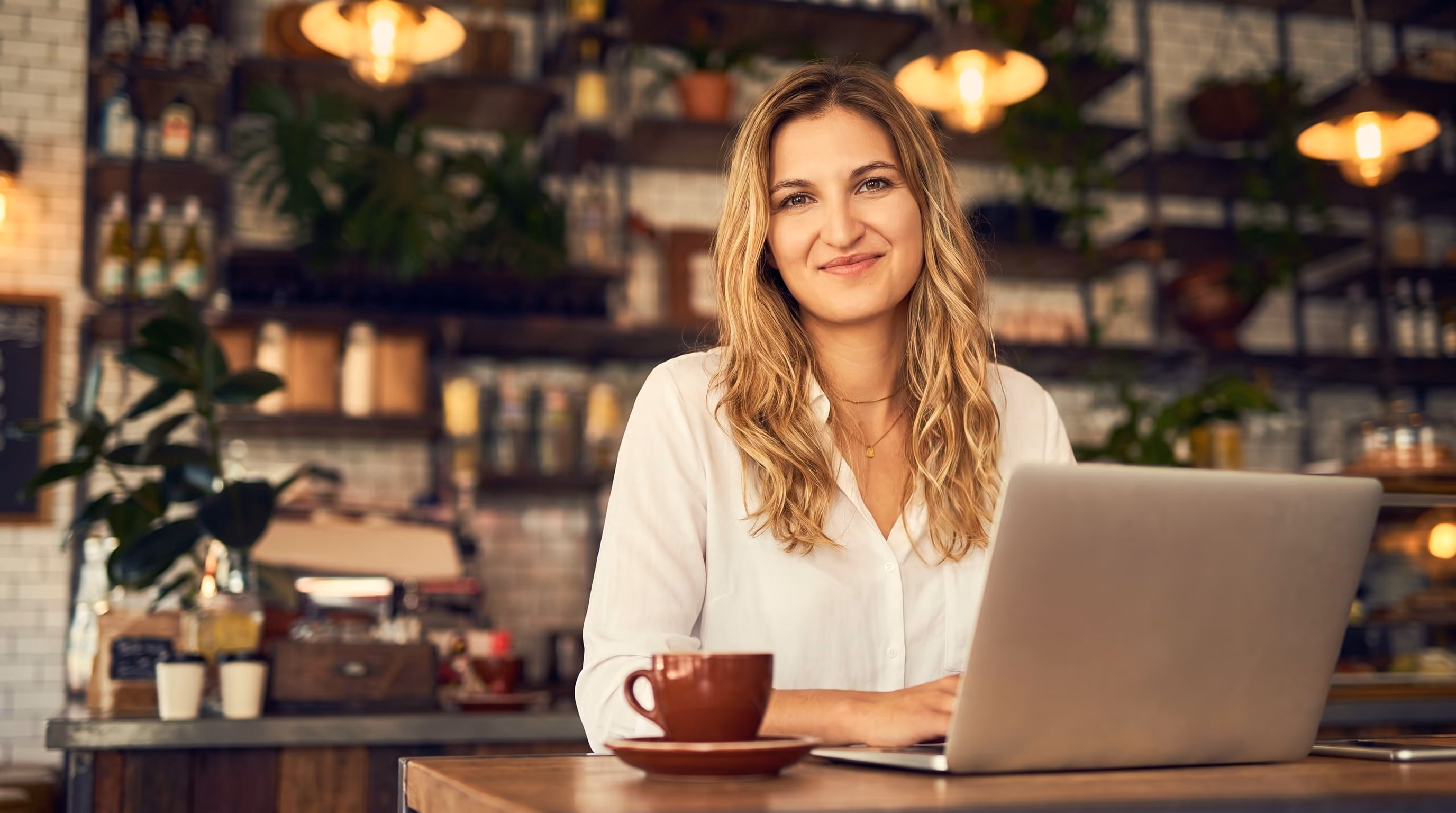 Lächelnde Frau sitzt im Café an Laptop mit Tasse Kaffee auf dem Tisch.