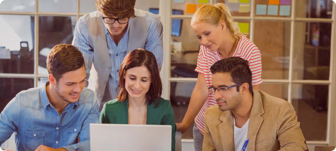 Five colleagues gathered around a laptop, collaboratively reviewing content in a modern office with glass walls and sticky notes.