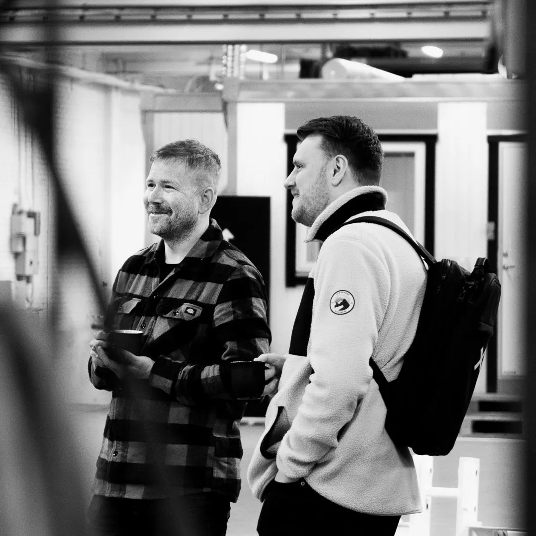 Två personer står och pratar med kaffekoppar i handen i en verkstadsmiljö – en avslappnad och genuin stund under arbetet.