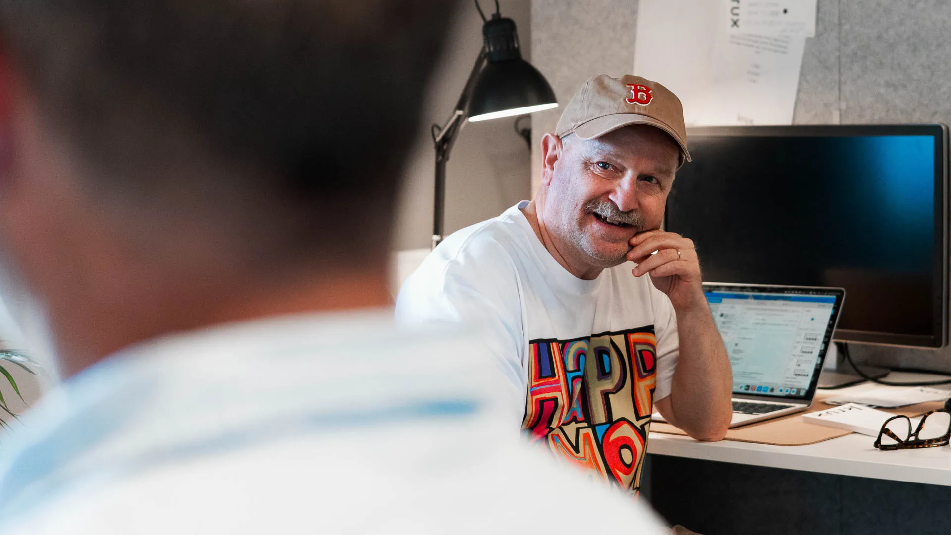 Leende person i keps sitter vid skrivbord med laptop och samtalar med en kollega på kontoret.