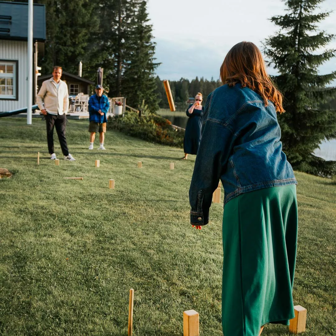 En grupp personer spelar kubb på en gräsmatta vid ett hus intill en sjö en sommarkväll.