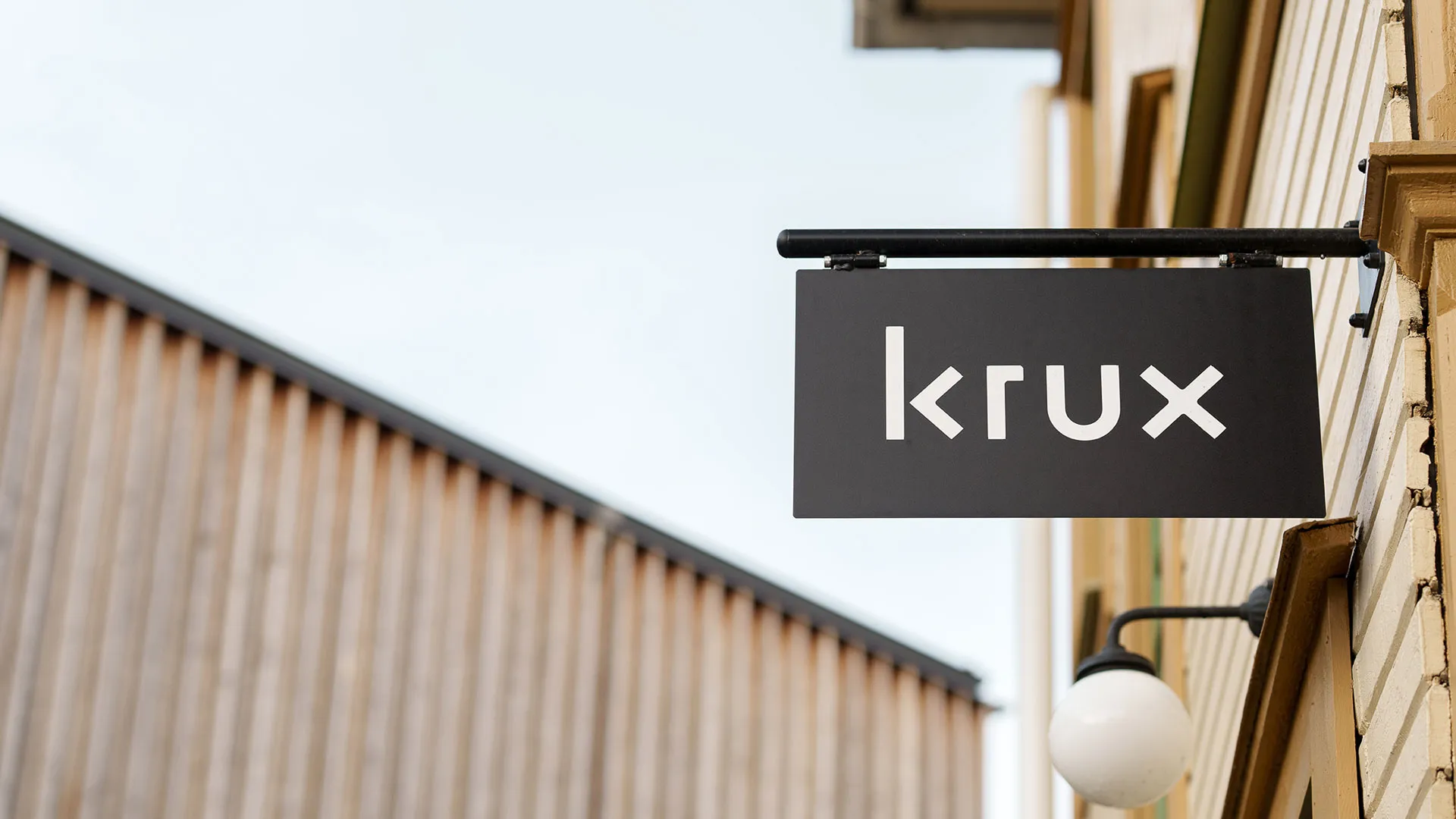 Svart skylt med vit text som visar logotypen “krux”, monterad på en husfasad i trä.