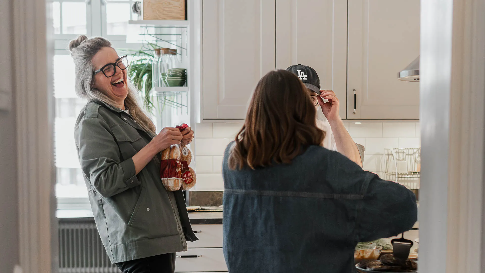 Tre personer står i ett kök och skrattar tillsammans, en av dem håller ett bröd i handen.