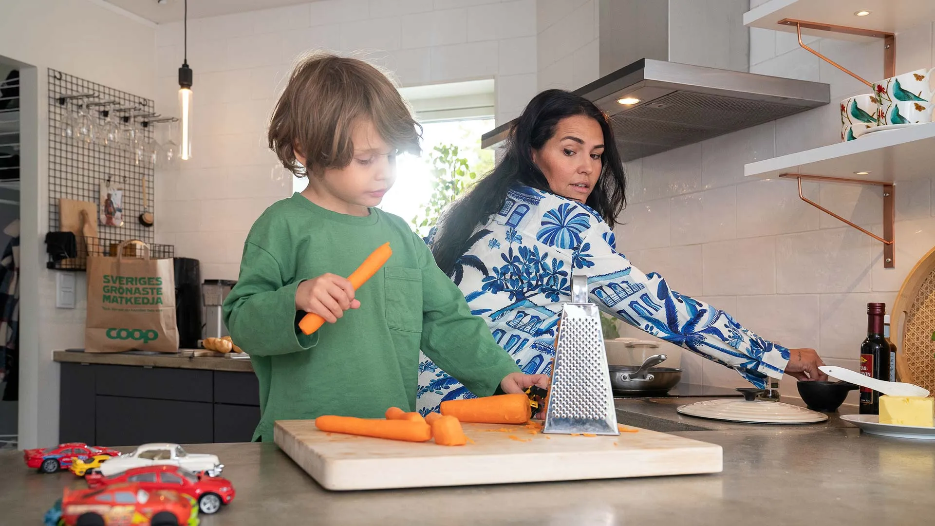 Barn och vuxen lagar mat tillsammans i ett kök, barnet håller en morot medan den vuxna förbereder något vid spisen.