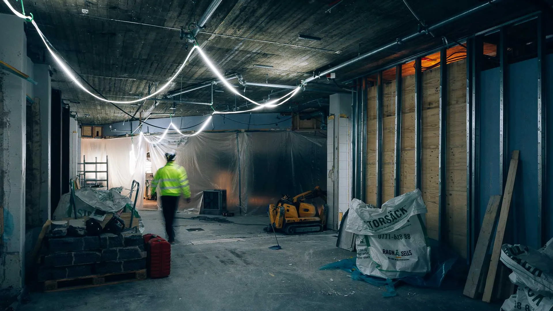 Person i varselkläder går genom en byggarbetsplats inomhus med verktyg, material och provisorisk belysning.