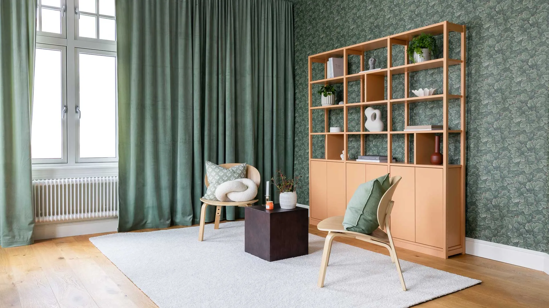 Ljust inreddt rum med två trästolar, grön vägg, gardiner och en hylla med dekorativa föremål.