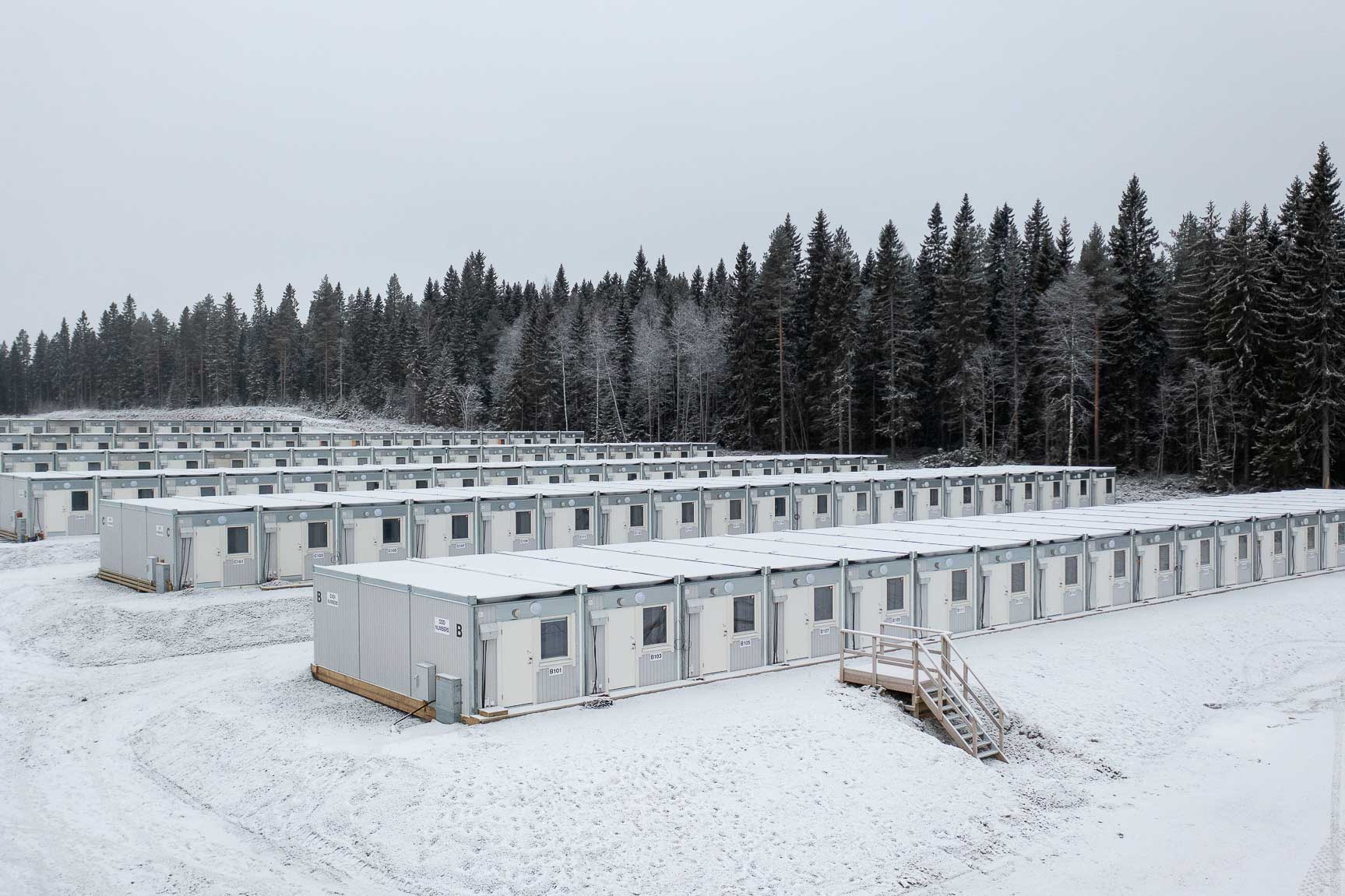 Rader av modulhus eller tillfälliga byggbaracker står uppställda på ett snötäckt område intill skog.