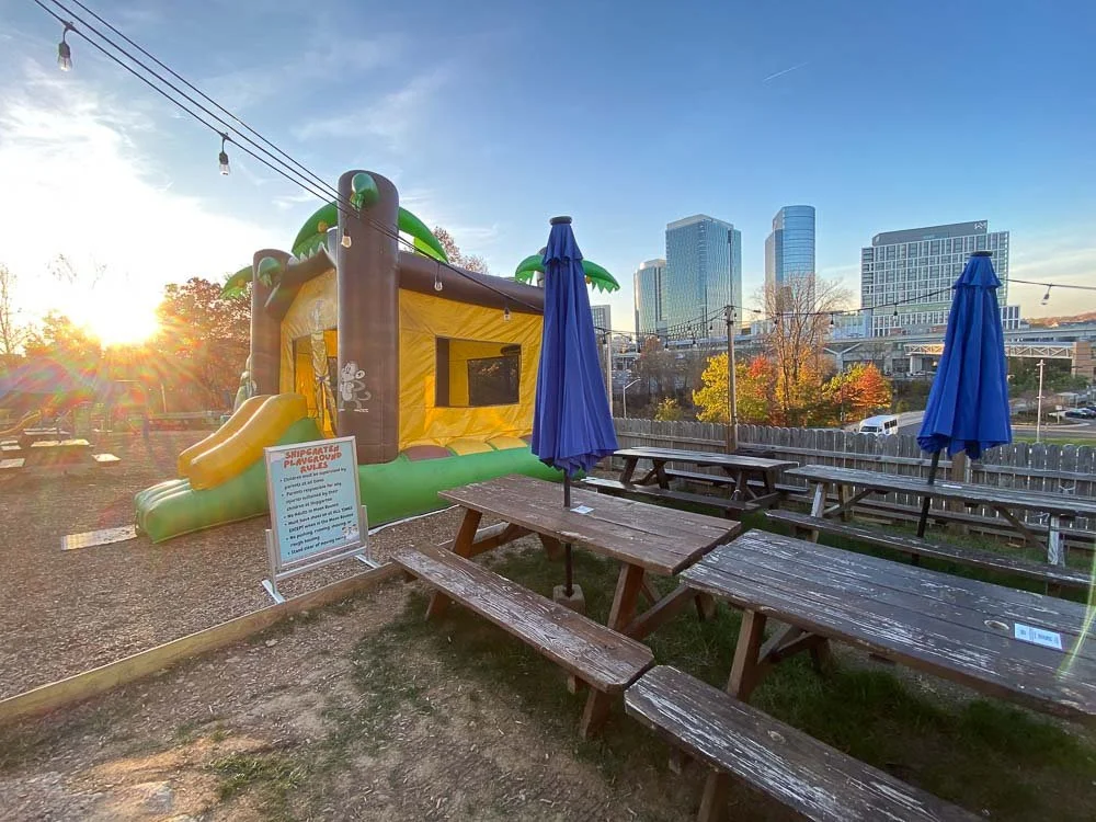 photo of picnic tables next to a bouncy castle, with string lights over head and office towers in the background