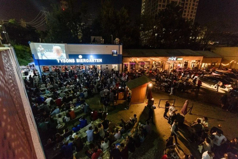 Aerial photo of the Tysons Biergarten at night. Several bars/restaurants face onto a patio area filled with people sitting at picnic tables and standing.