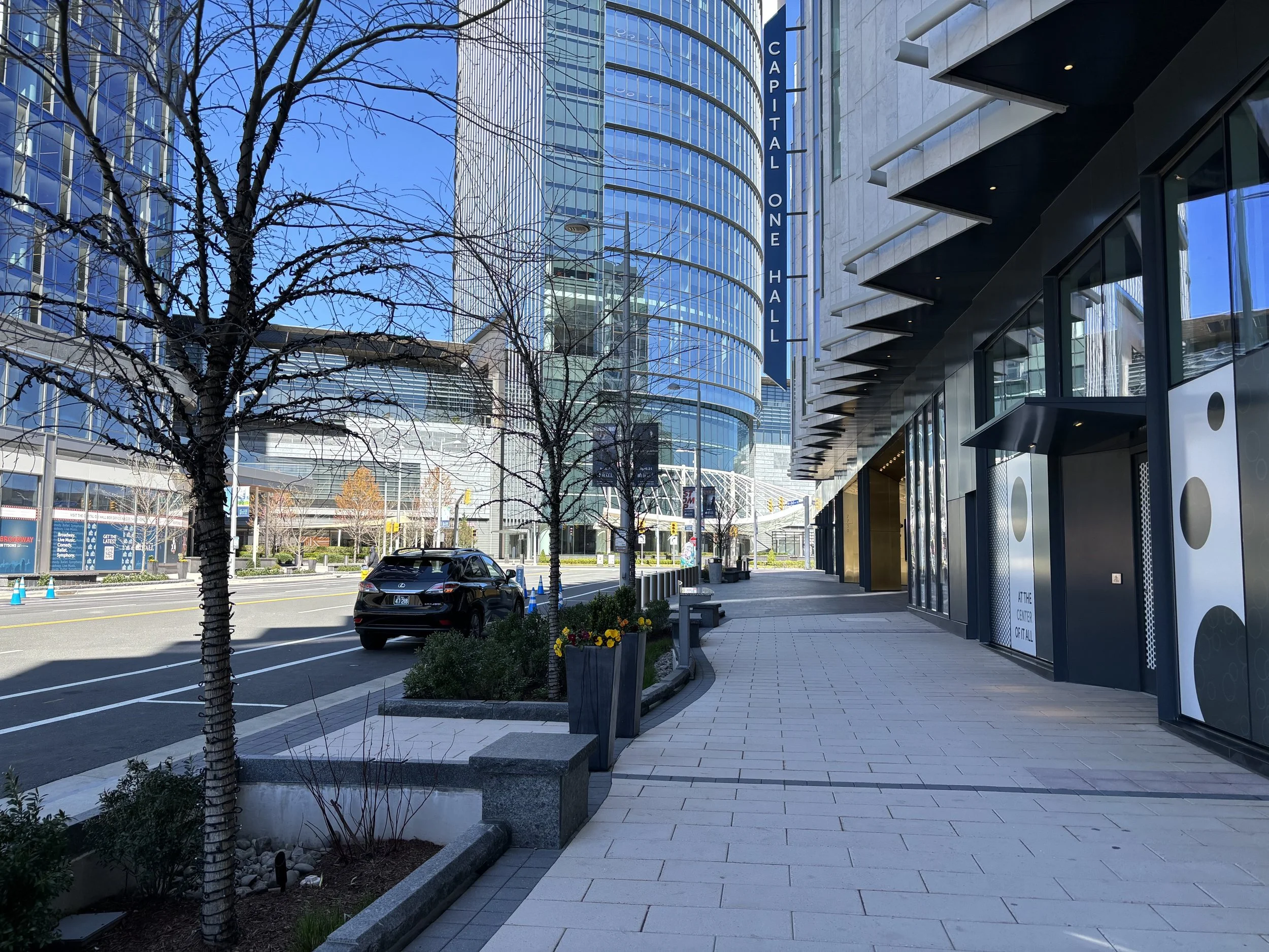 Photo of new development in Tysons from street level. There is a wide sidewalk buffered from the road with planter beds. The street is lined with tall modern buildings