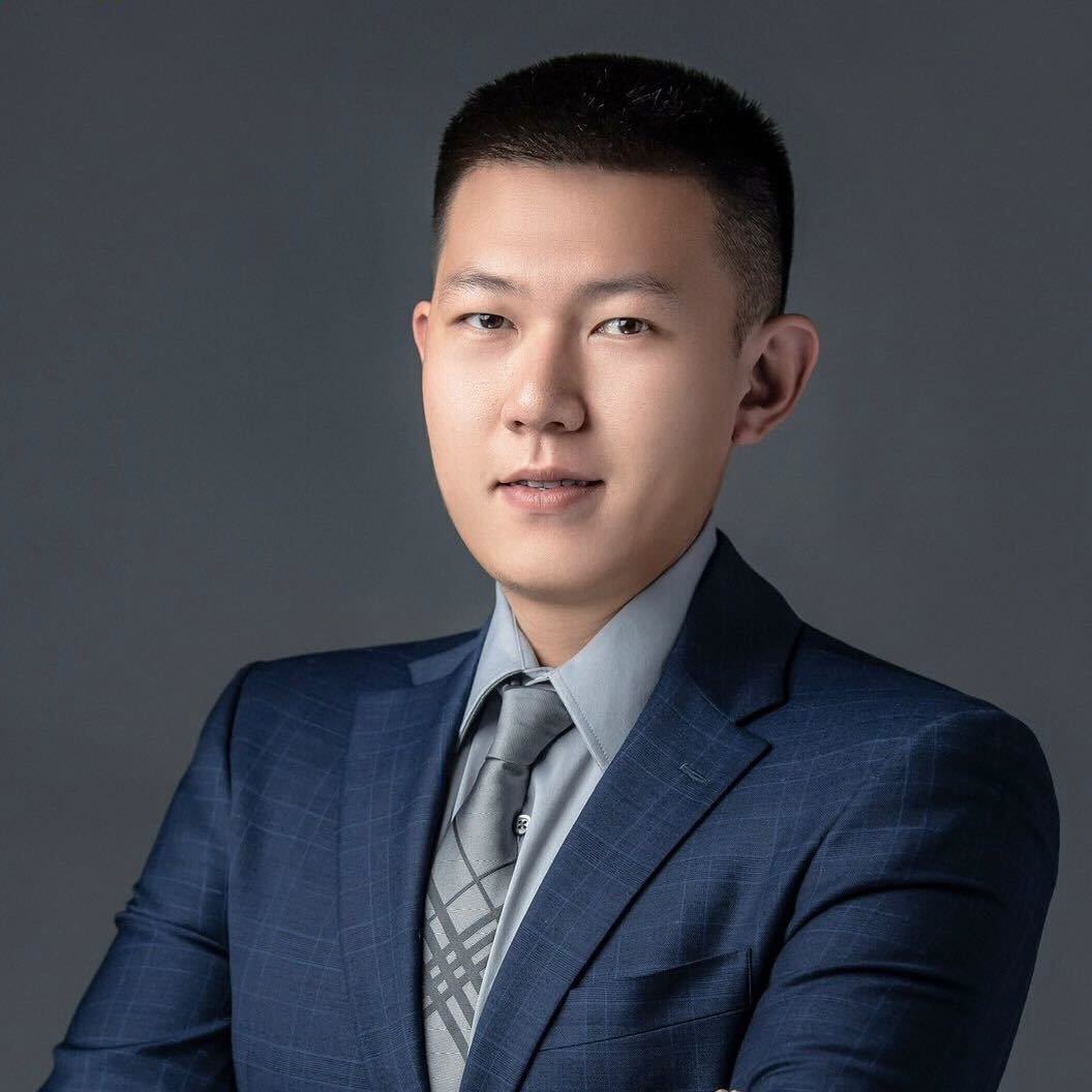 Portrait of a young man wearing a blue suit, gray shirt, and patterned gray tie against a dark gray background.