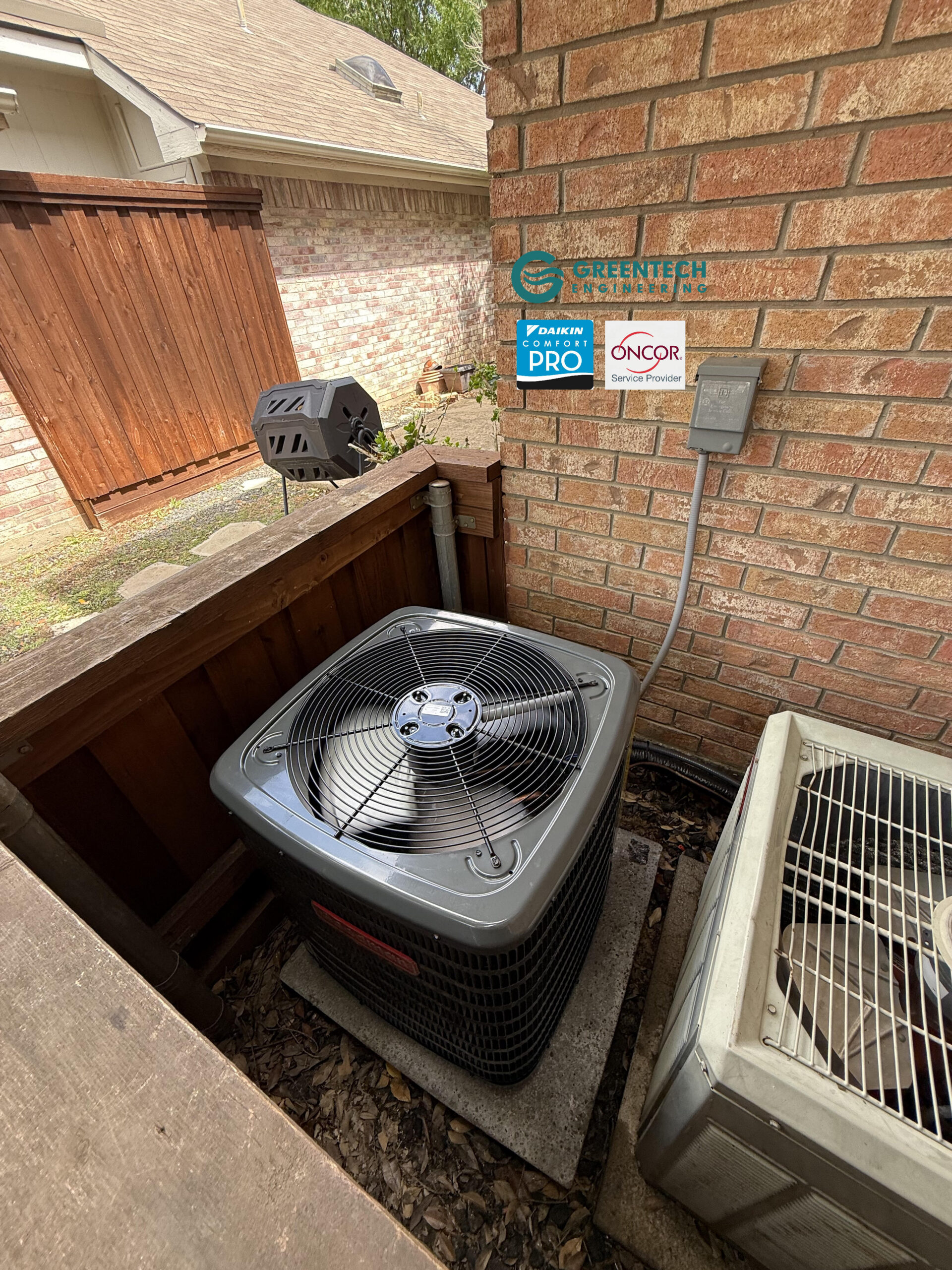 A newly installed, dark gray Goodman 2-Ton AC condenser sitting securely on an equipment pad on a second-story balcony of a Dallas home, with a new electrical disconnect box on the brick wall.